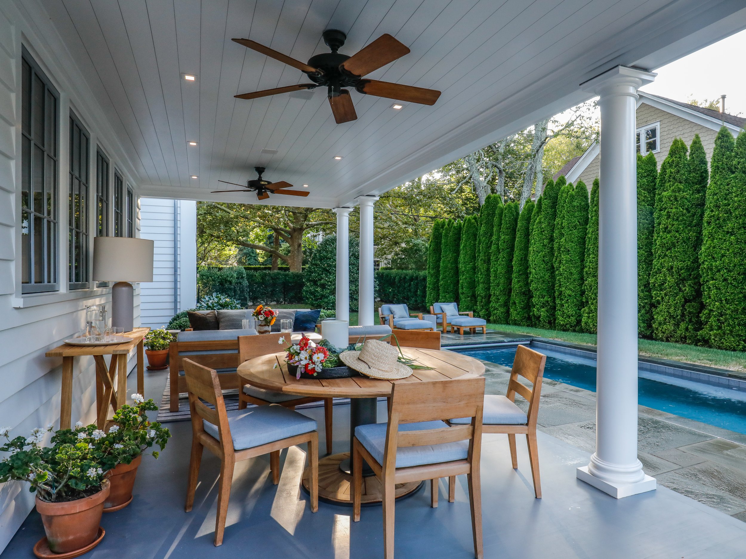 Covered patio with wooden dining table, chairs, potted plants, ceiling fans, and a swimming pool with lounge chairs in a backyard with tall green hedges. Construction in Hamptons New York
