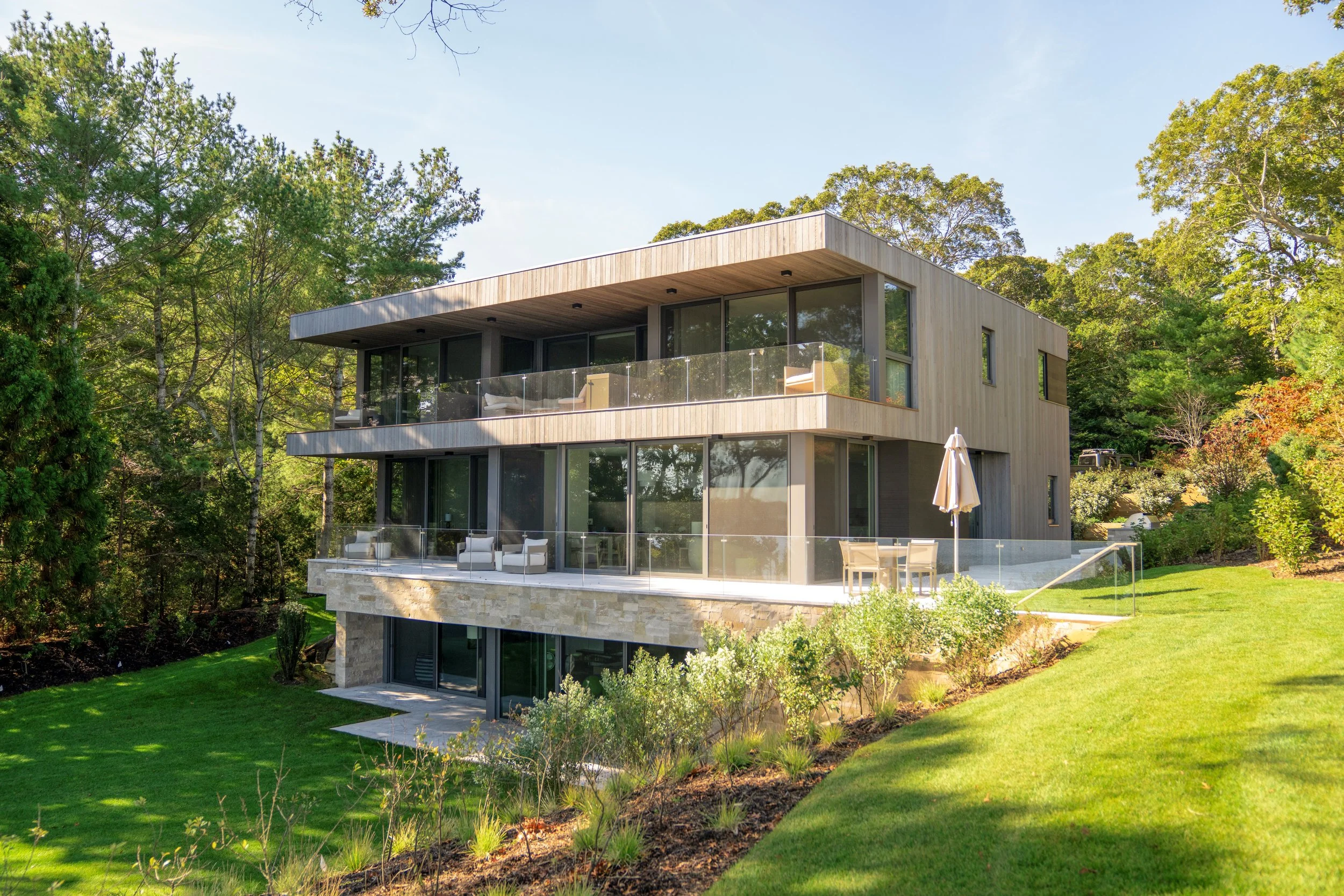 Modern multi-story house with large glass windows and balconies, surrounded by green lawn and trees.