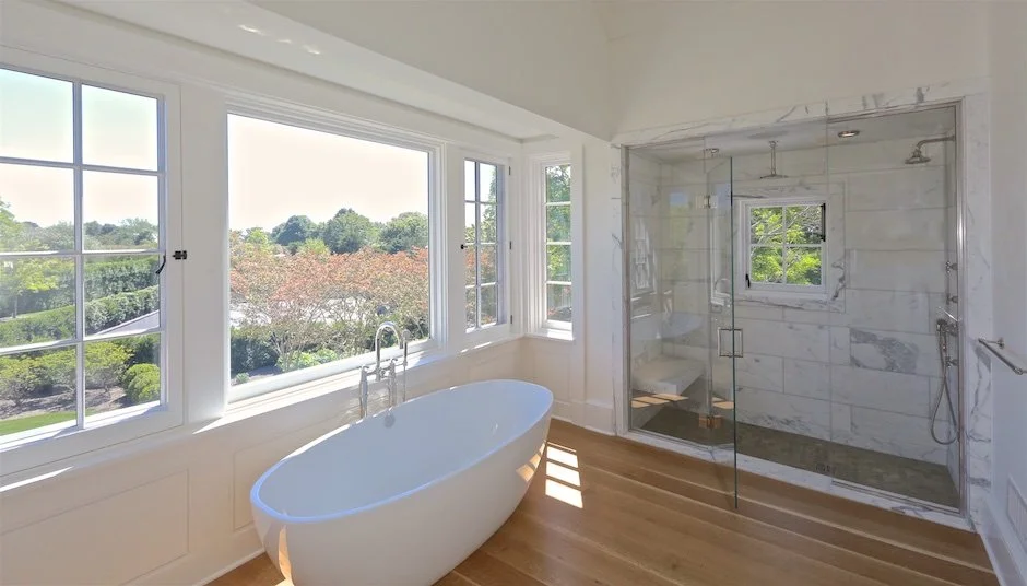 Bathroom with white freestanding bathtub beneath large windows, glass-enclosed shower with marble walls, and wooden flooring, overlooking a green landscape. Montauk New York