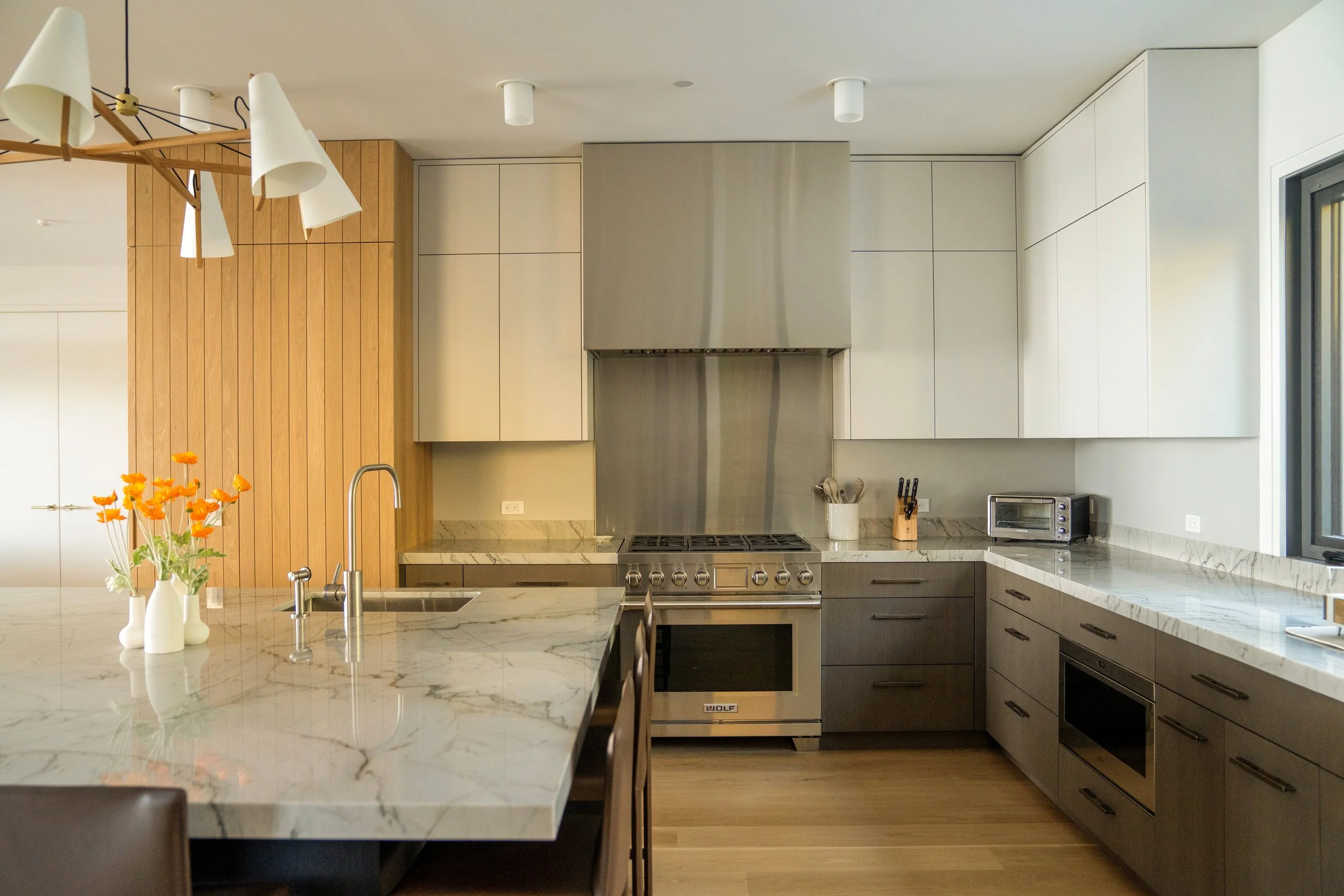 Modern kitchen with marble countertops, stainless steel appliances, and white cabinetry. There is a wooden wall accent, a vase with orange flowers, and a dining area with chairs.