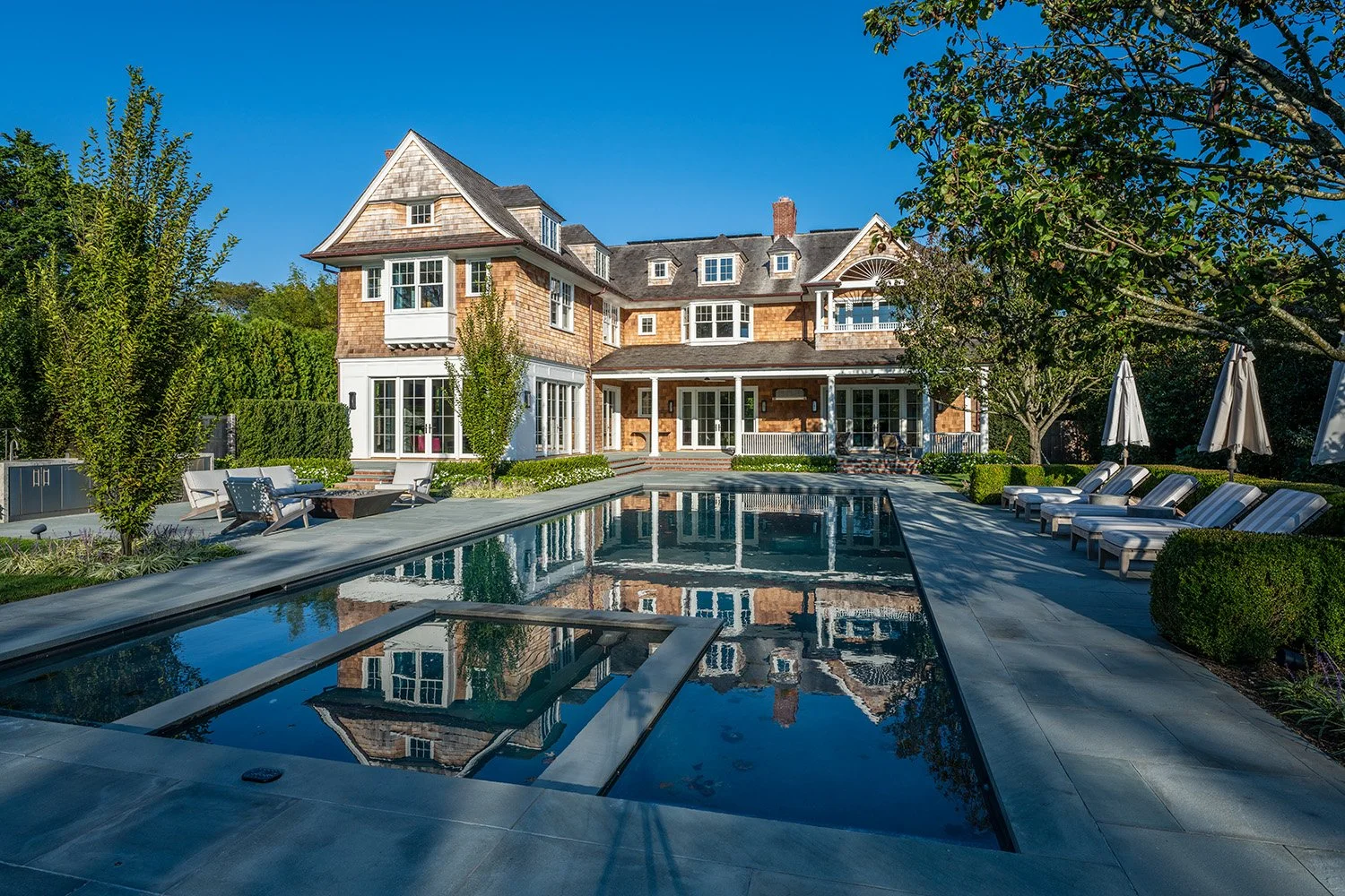 Large upscale house with a swimming pool in the backyard, landscaped with trees and lounge chairs, under a clear blue sky.
