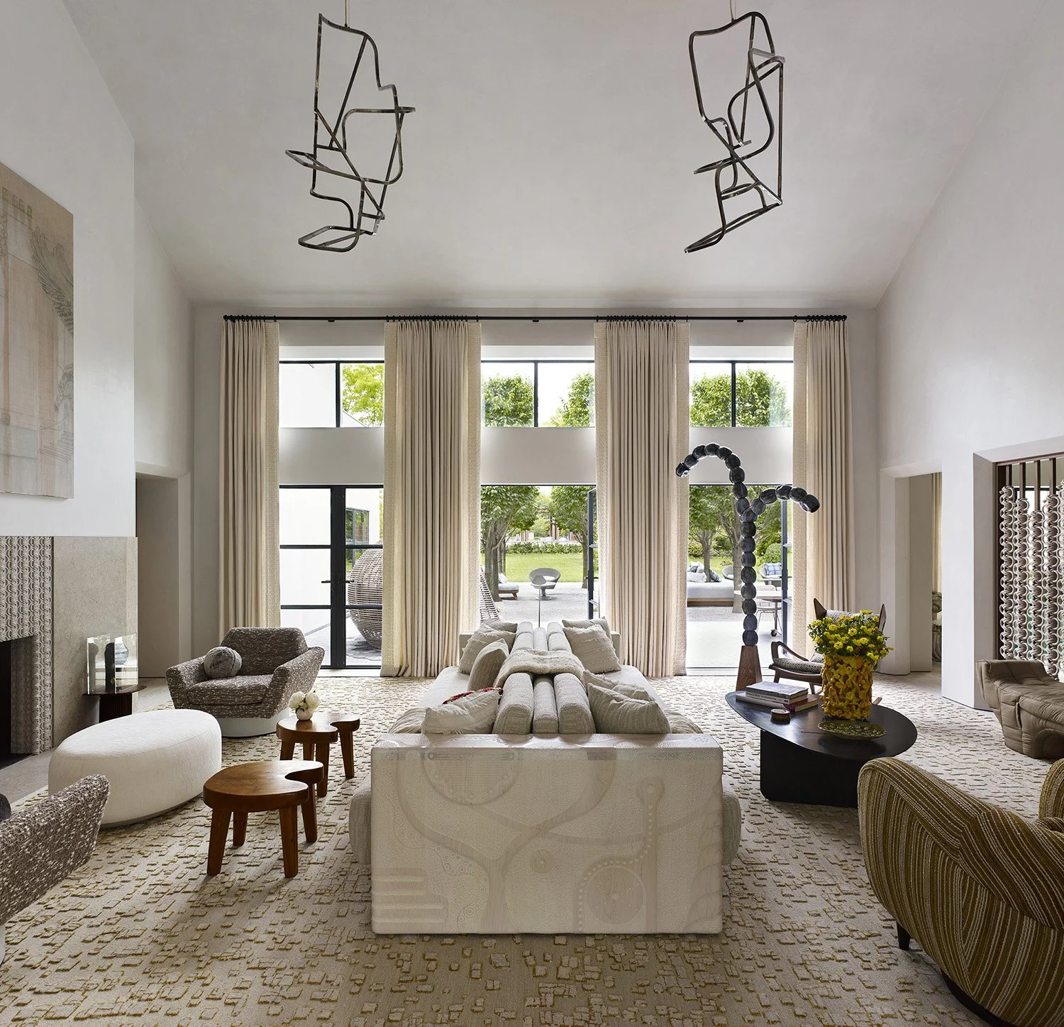 Modern living room with large windows, beige curtains, and various furniture pieces, including a sofa, armchair, coffee table, and decorative sculptures.