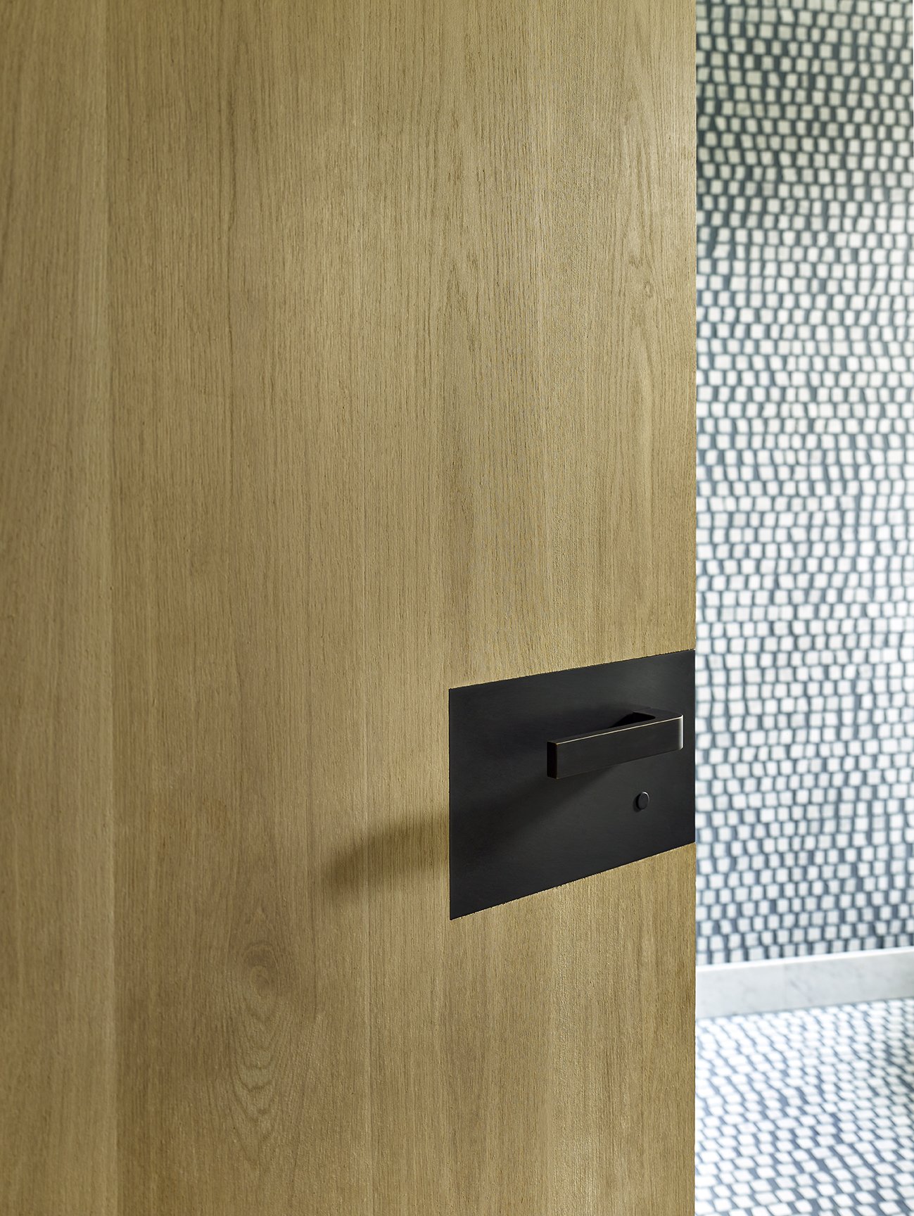 Close-up of a wooden door with a black rectangular door handle on the left side, and a patterned wall with small square tiles in the background.