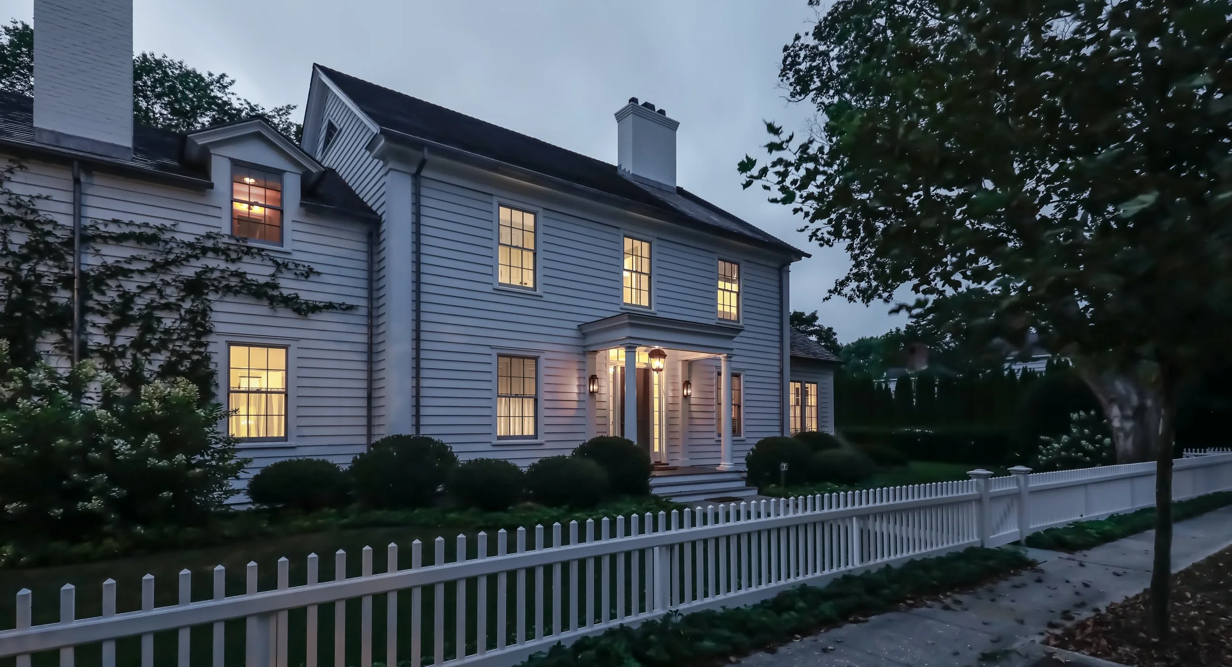 White two-story house with lit windows at dusk, surrounded by a white picket fence and greenery. Construction in Hamptons New York