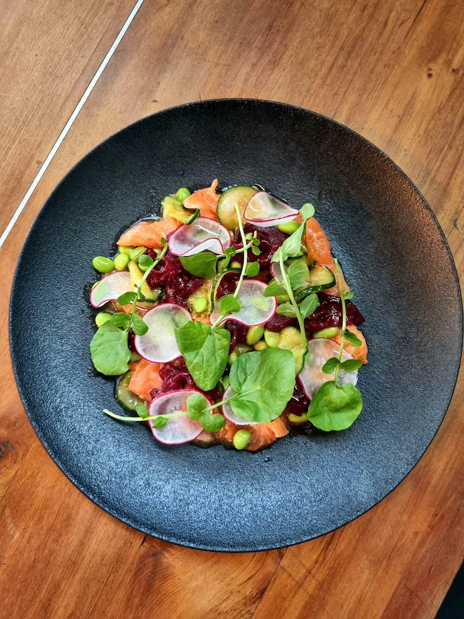 Salad with radish slices, cucumber, salmon, mixed greens, and edamame, served on a black plate on a wooden table.
