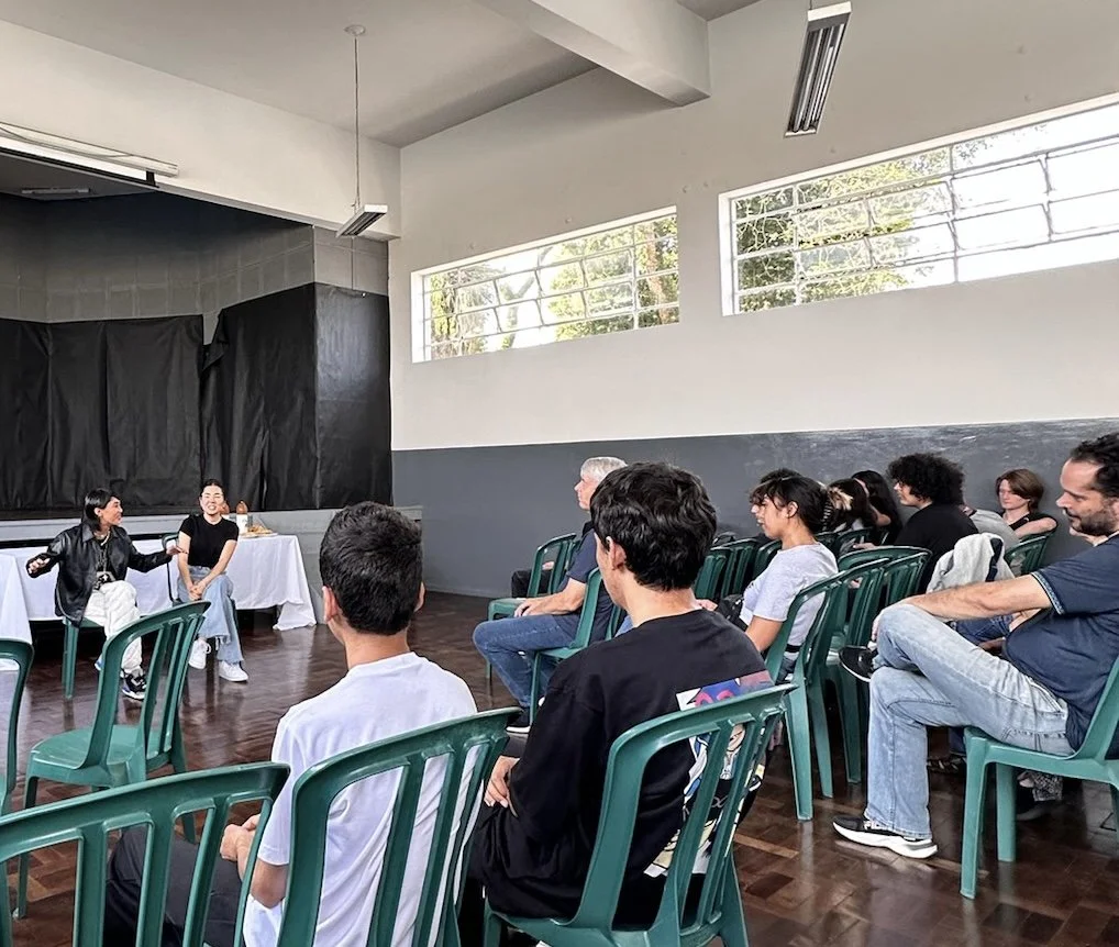 A small group of people seated in green chairs watching a small stage where two women are sitting and talking.