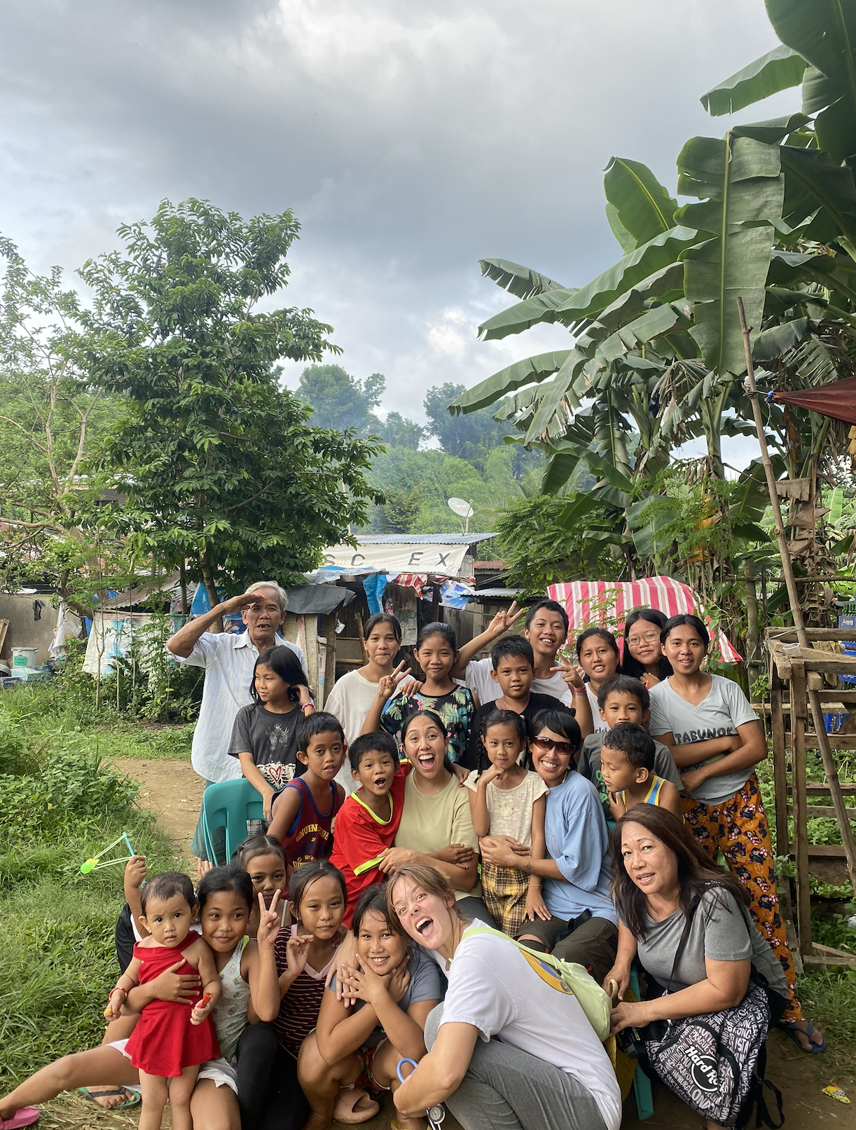 A group of smiling children, women, and an elderly man posing outdoors in a rural area with lush green trees and houses in the background, some making peace signs or salutes.
