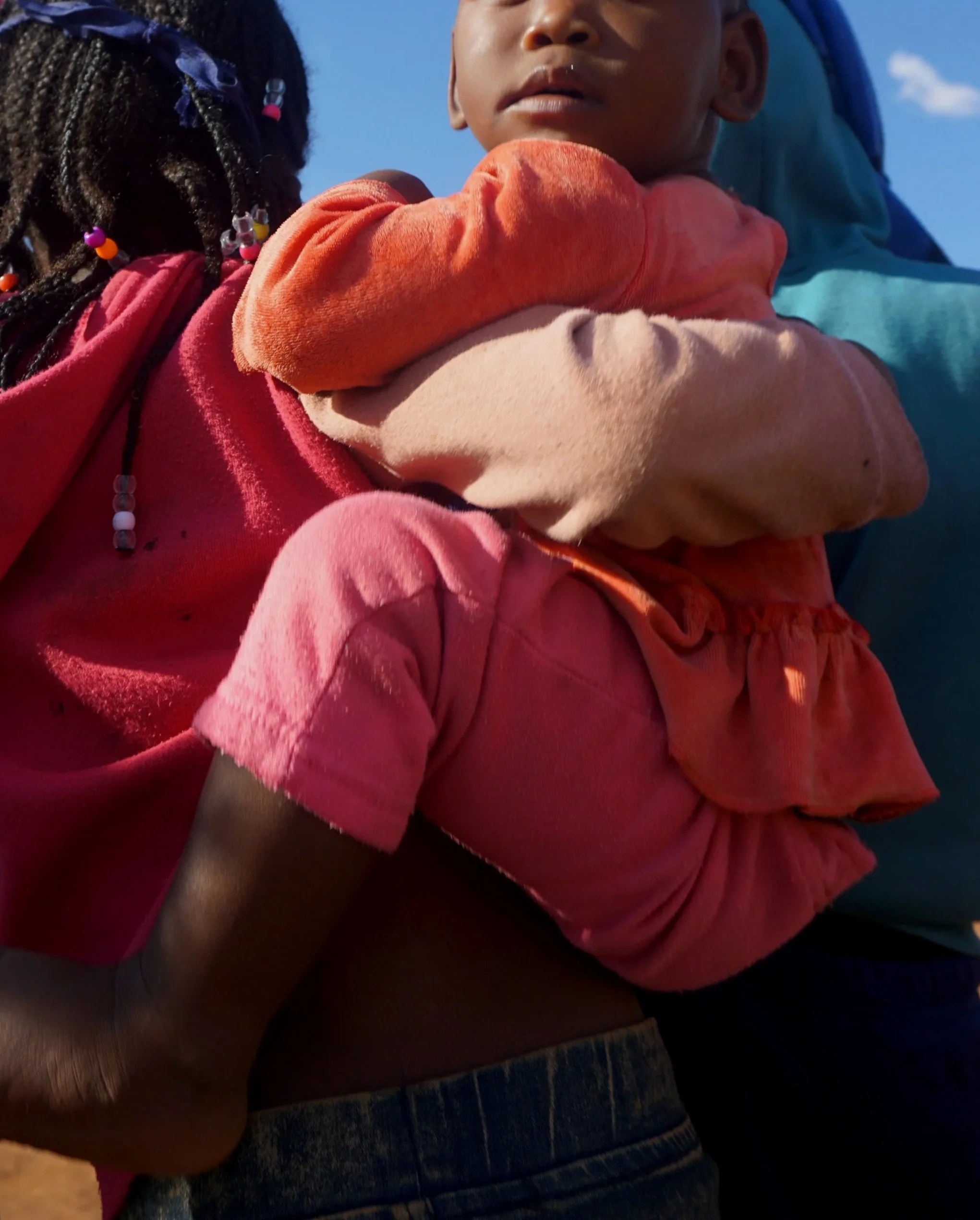 Close-up of a person holding a child with the child's face partially visible, wearing colorful clothing and a blue hoodie, against a clear blue sky background.
