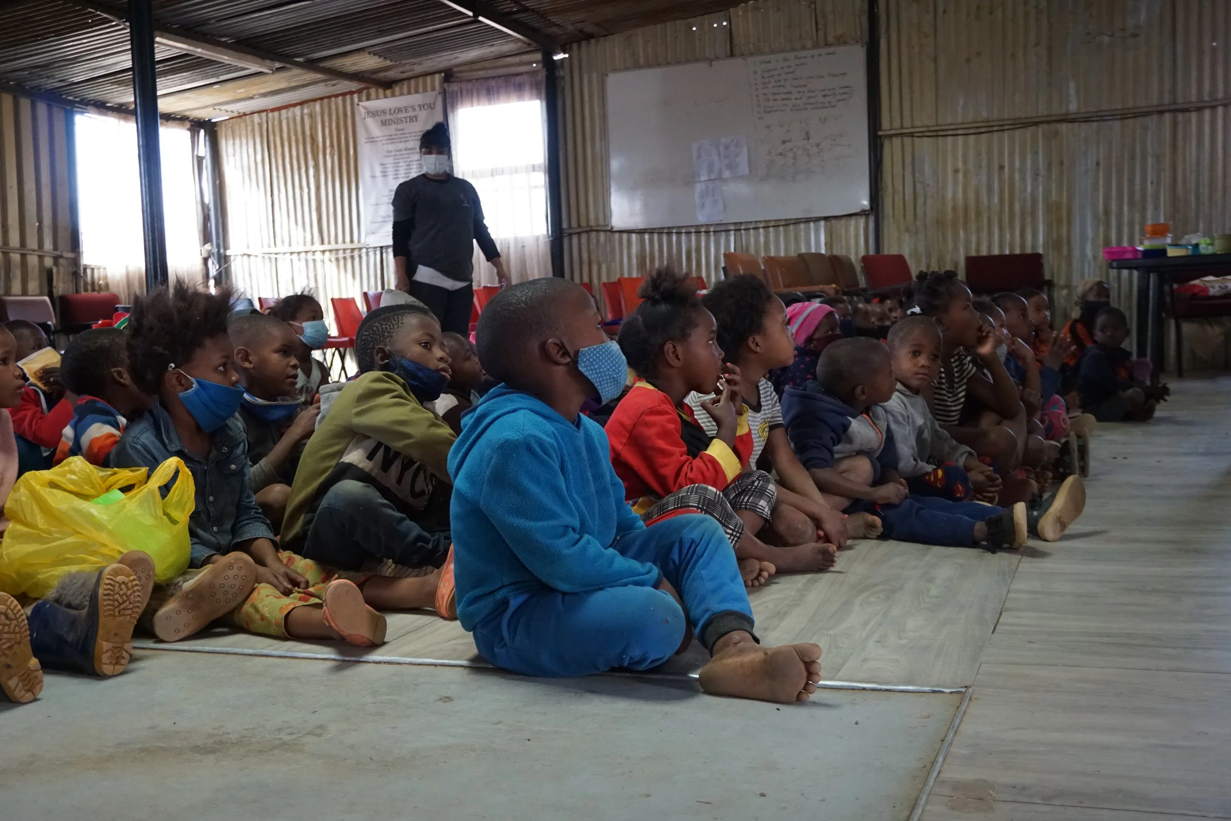 Group of children sitting on the floor inside a rustic wooden building, attentively facing forward, with a woman standing at the back near a whiteboard and windows, some children wearing face masks.