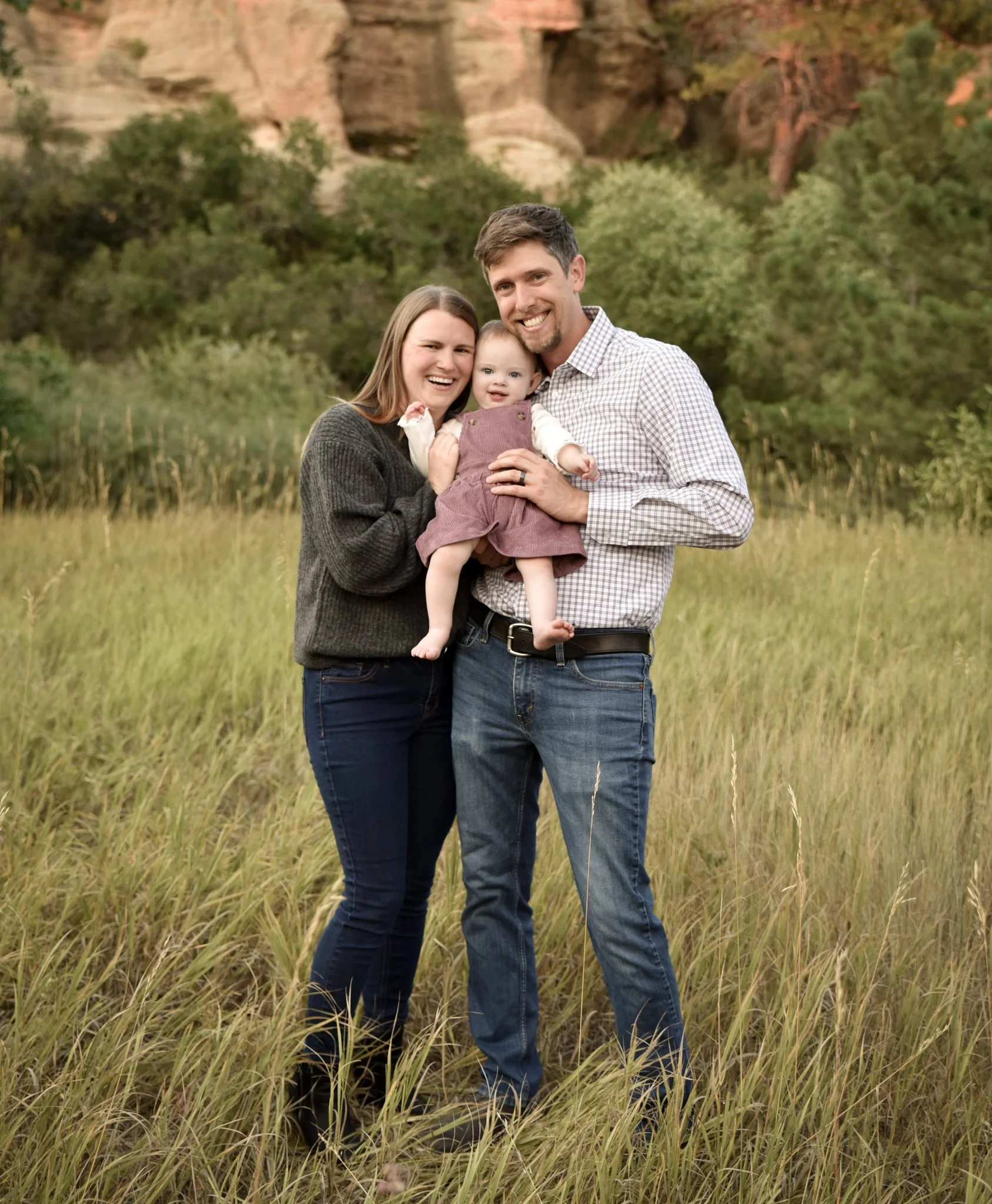A family of three outdoors during autumn, with trees changing color in the background, the mother and father smiling while holding their young child between them.