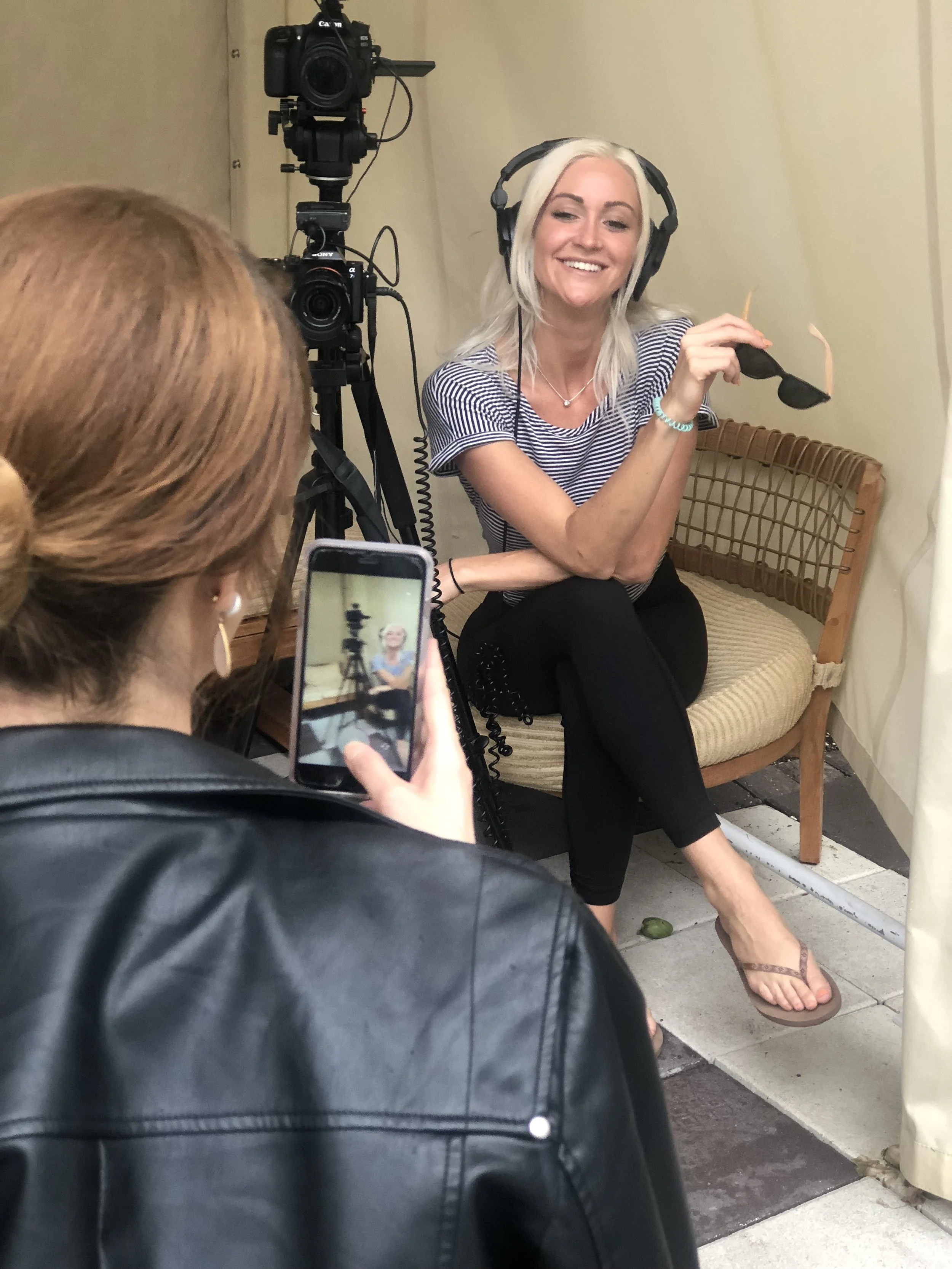 A woman with blonde hair, wearing headphones, a striped shirt, and black pants, is sitting on a bench and smiling while being filmed. Another person, with red hair and wearing a leather jacket, is recording her with a phone. A camera on a tripod is p