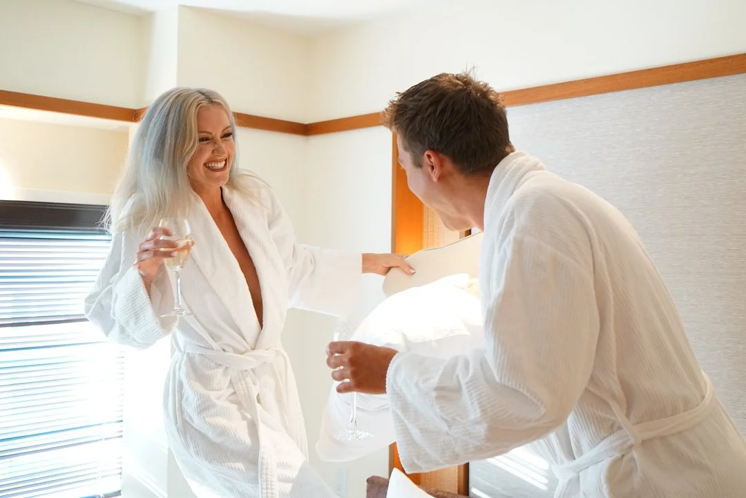 A woman and a man wearing white bathrobes celebrating in a hotel room, holding glasses of champagne, smiling and laughing.