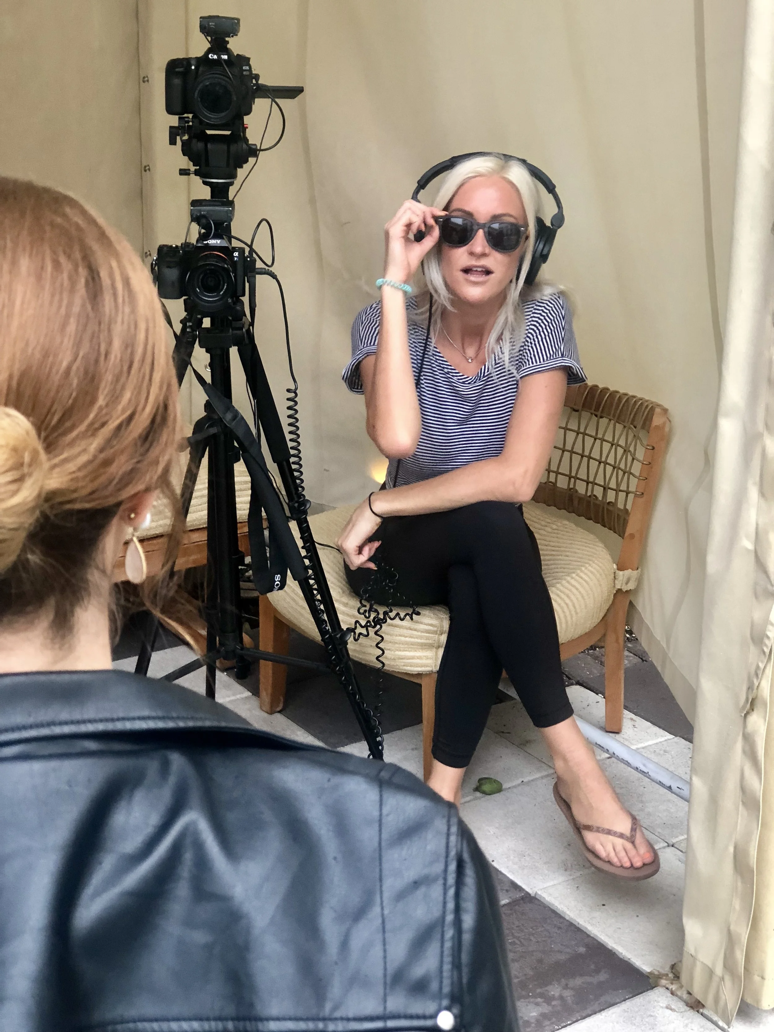A woman with blonde hair wearing sunglasses, a striped shirt, and black pants is sitting on a wooden chair with a cushioned seat, speaking to someone in front of her. She is wearing headphones and adjusting her sunglasses. A camera on a tripod is poi