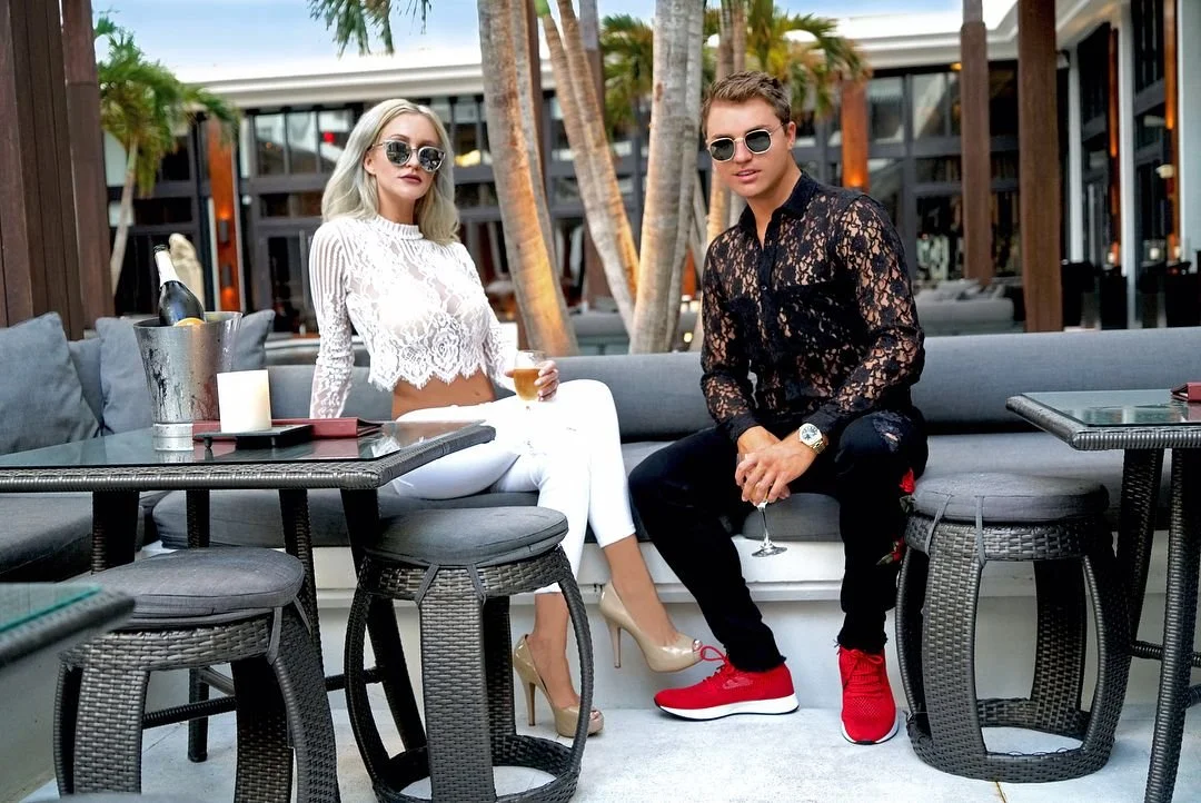 A stylish man and woman sitting outdoors on a modern patio, holding drinks. The woman is wearing a white lace crop top, white pants, beige high heels, and sunglasses. The man is dressed in a black lace shirt, black pants, red sneakers, and sunglasses