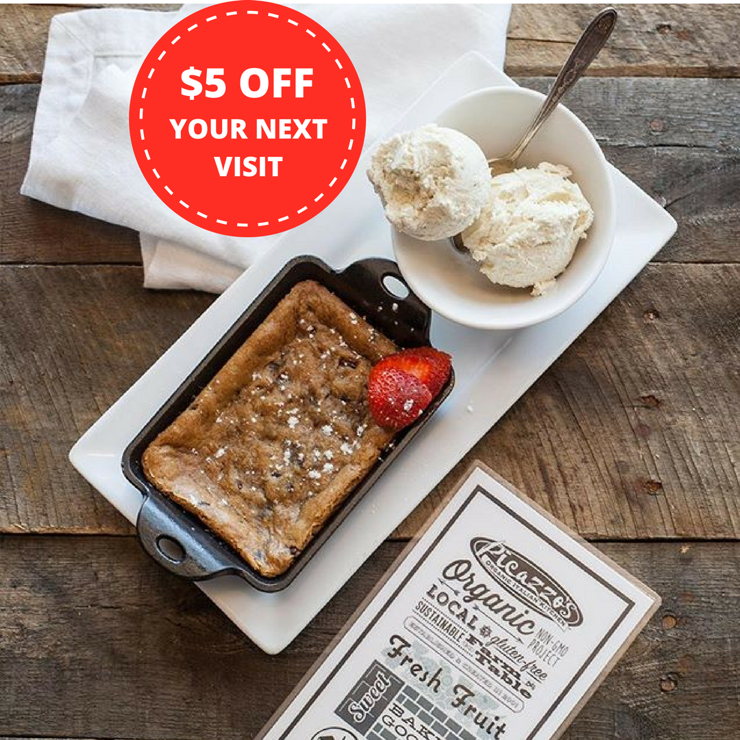 A dessert plate with a small cast iron skillet of caramel apple crumble, a scoop of vanilla ice cream in a white bowl with a spoon, a sliced strawberry, and a promotional flyer offering five dollars off a next visit, all on a rustic wooden table.