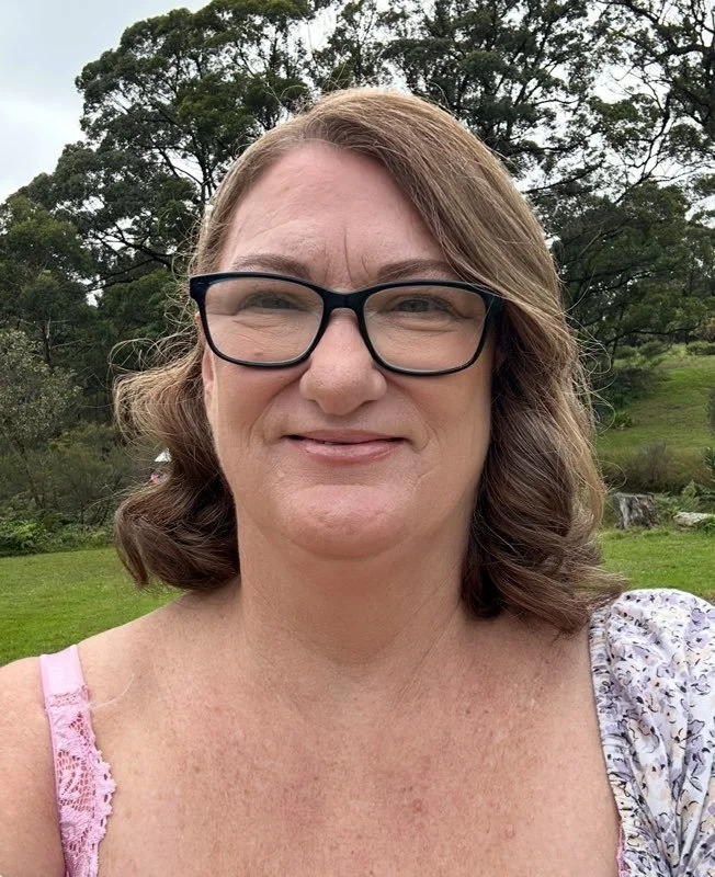 A woman with shoulder-length brown hair, wearing black glasses and a light-coloured, floral-patterned top, smiling outdoors with greenery and trees in the background. The woman is Karen James, a Reiki Healer, owner of Insightful Vibrations.
