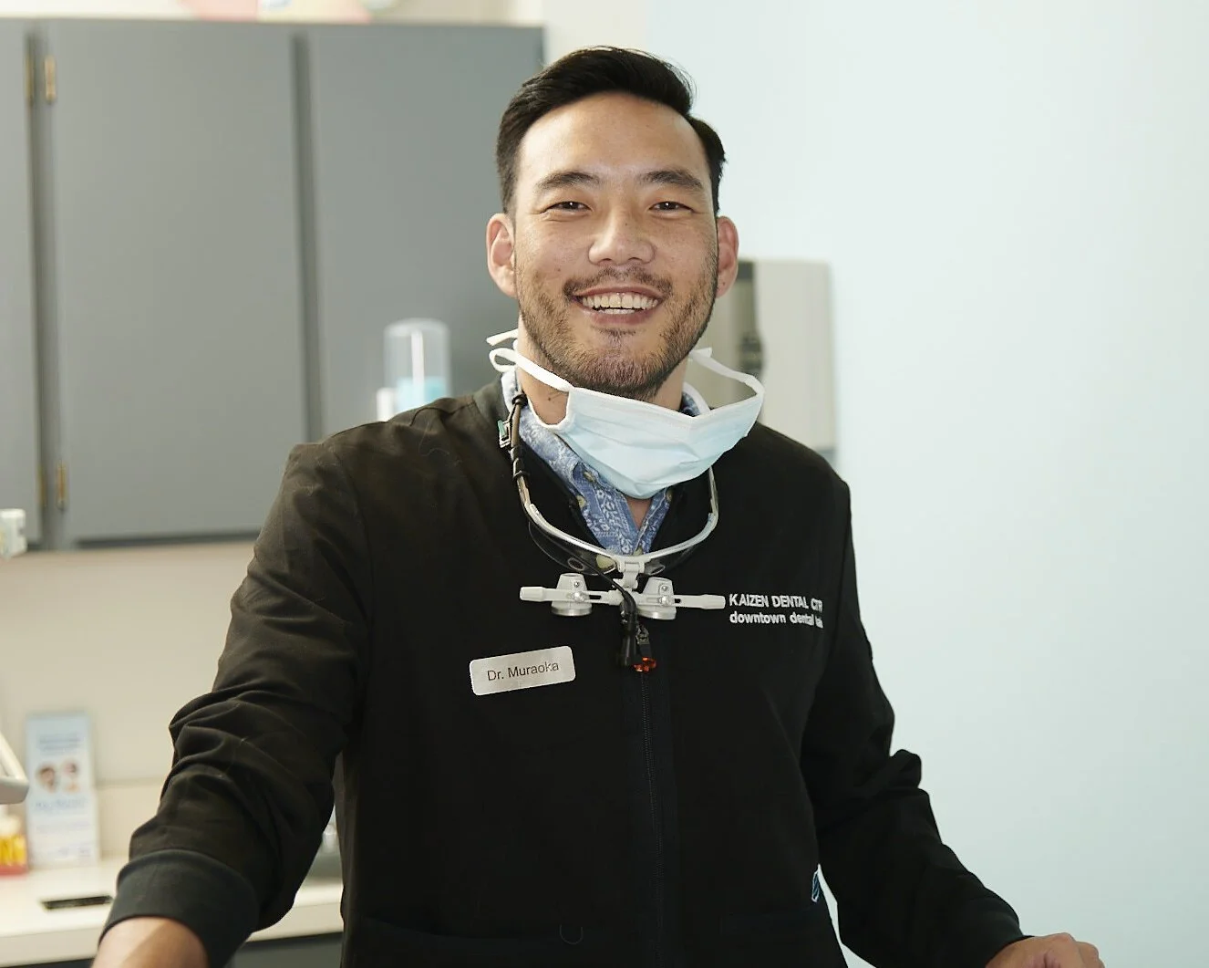 A smiling male dentist in a black coat with a name tag, standing in a dental office with a face mask lowered on his chin.