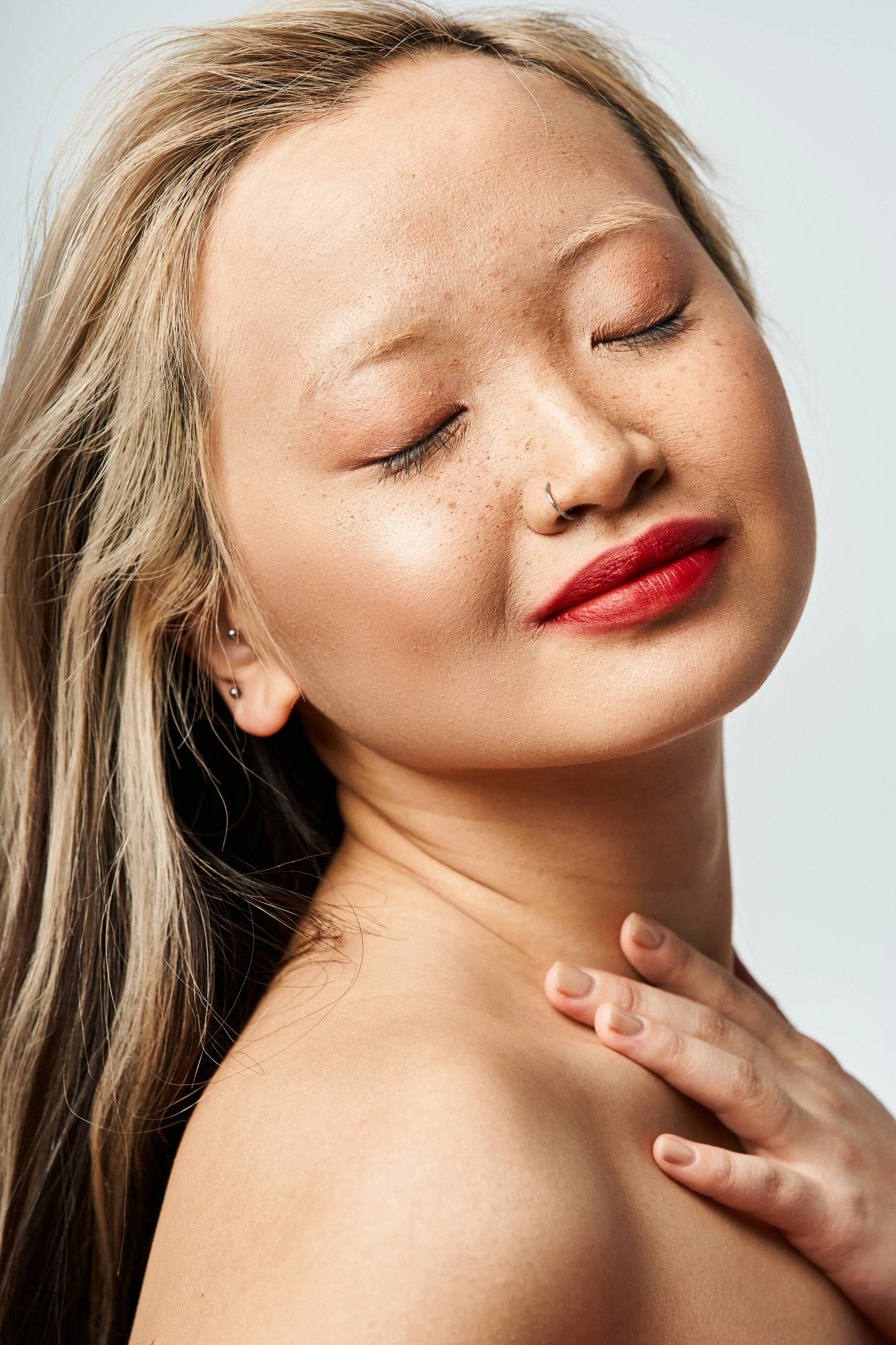 Close-up of a woman with blonde hair, makeup, a nose ring, and multiple earrings, with closed eyes and hand on her shoulder.