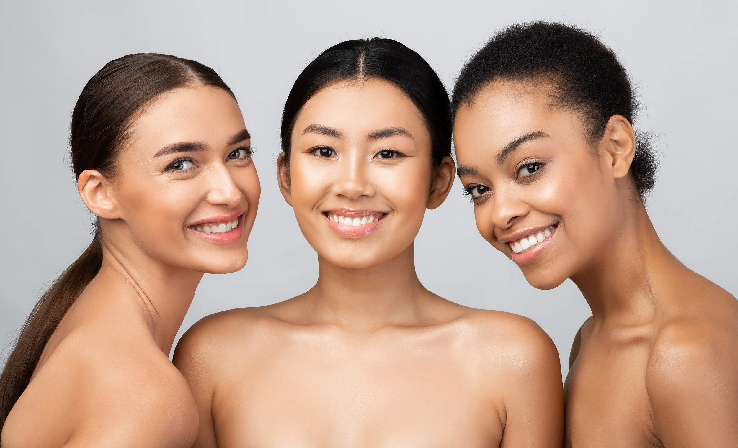 Three women with different skin tones smiling and looking at the camera, standing close together against a plain background.