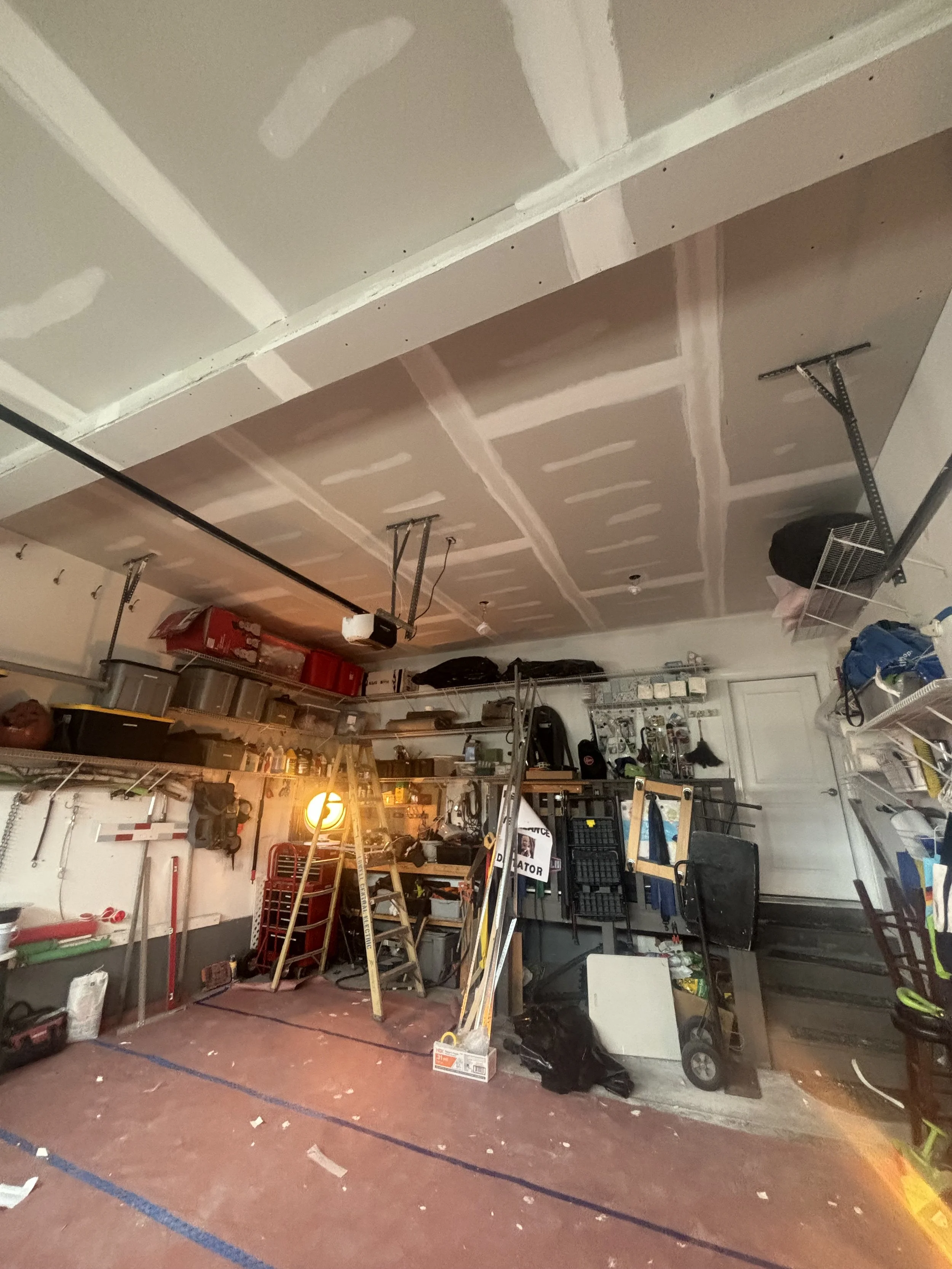 A cluttered garage with tools, storage shelves, and various items, including a ladder, boxes, and a trash bag, with a ceiling that is being renovated or still under construction.