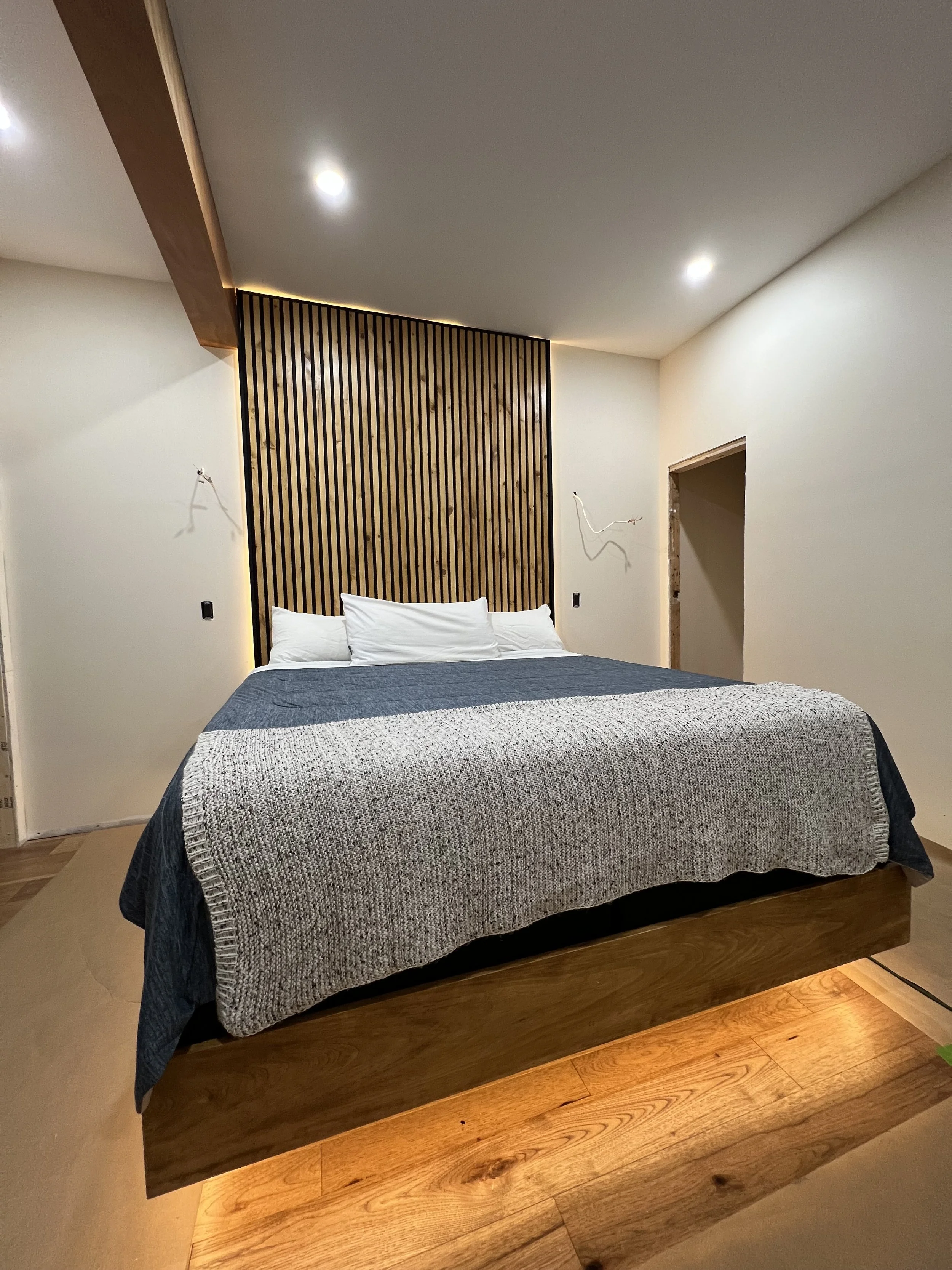 Modern bedroom with wooden bed frame, gray bedding, white pillows, and a wooden slat accent wall behind the bed, lit with recessed ceiling lights and warm floor lighting.