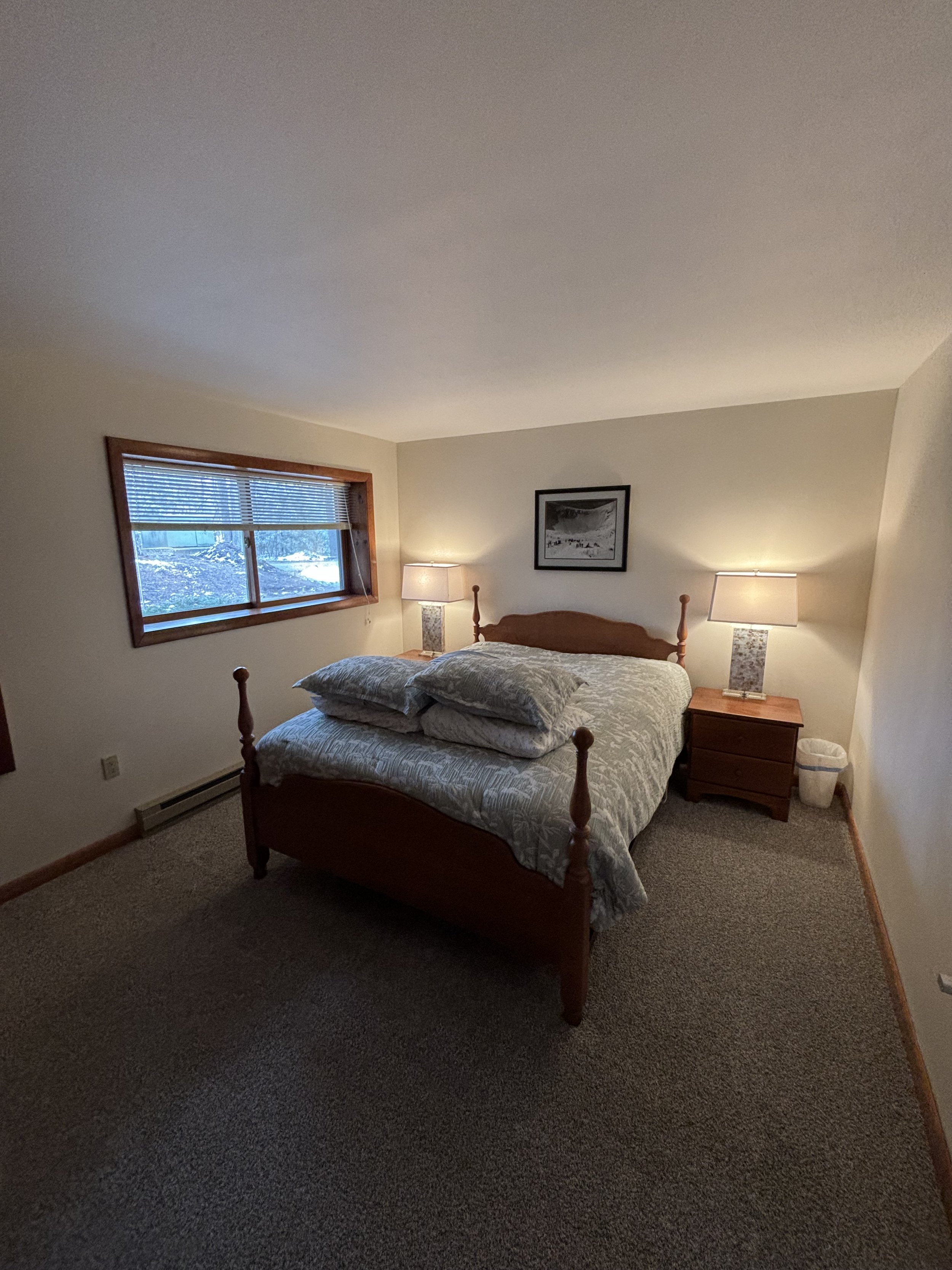 A bedroom with a wooden bed, gray patterned bedspread, and two pillows. There are two matching bedside tables with lamps, a framed black-and-white picture on the wall, a window with blinds, and a trash bin on the carpeted floor.