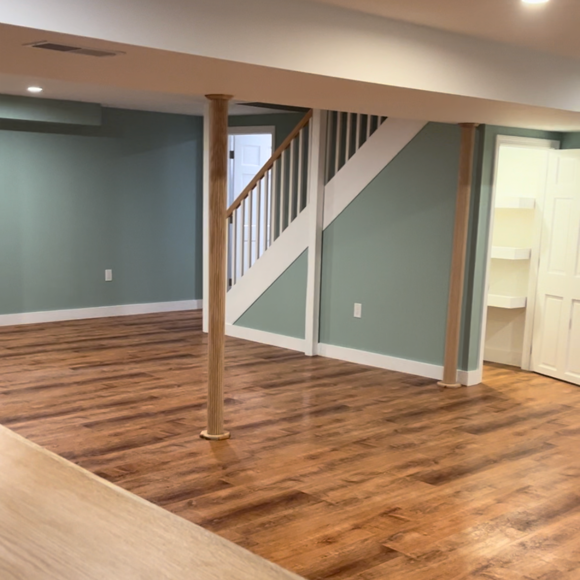 Empty living room with hardwood flooring, teal walls, staircase with white and wood railing, and a small white closet or storage area with open shelves.