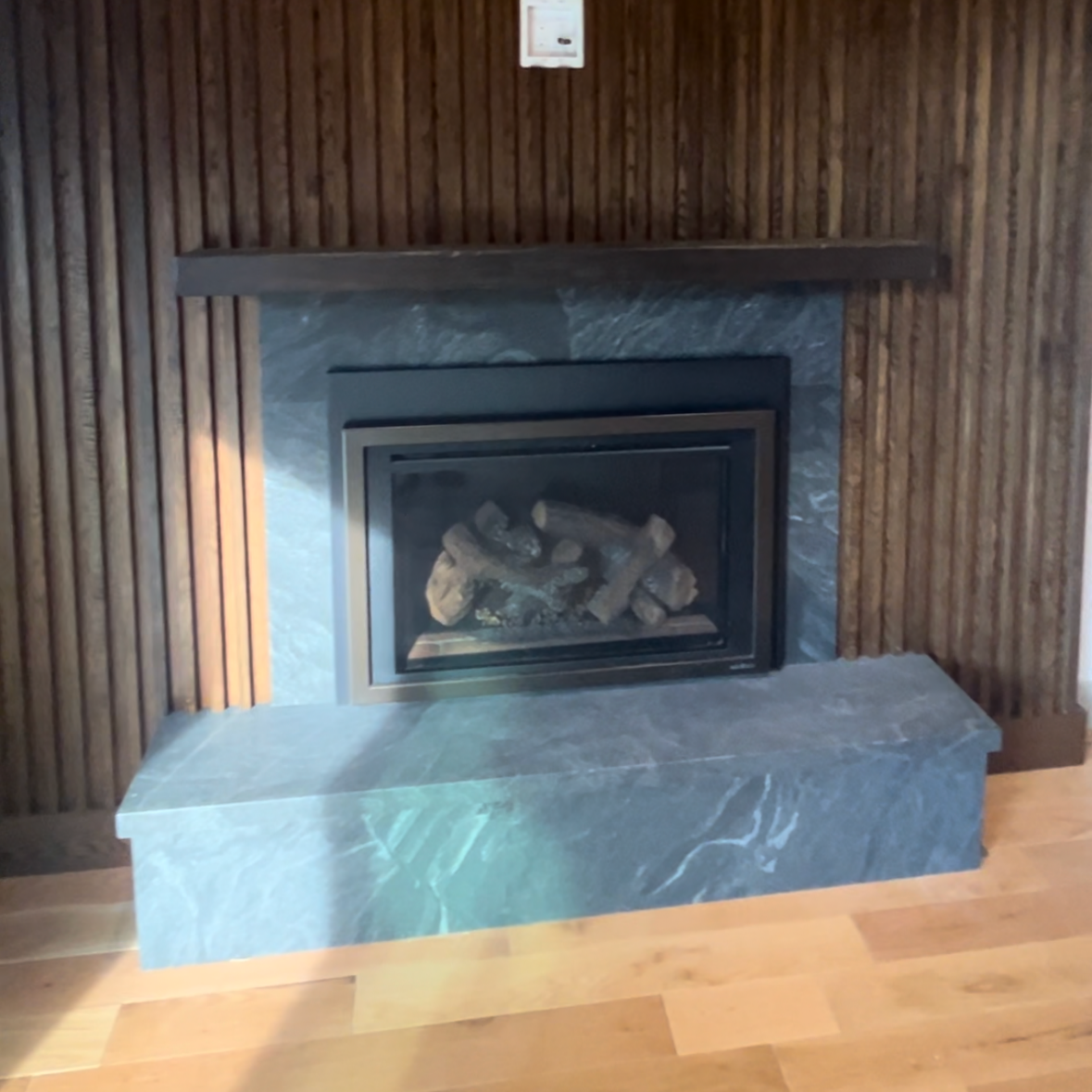 A modern fireplace with a black metal frame, installed in a wall with vertical wooden paneling, on a blue-gray marble hearth, on a wooden floor.