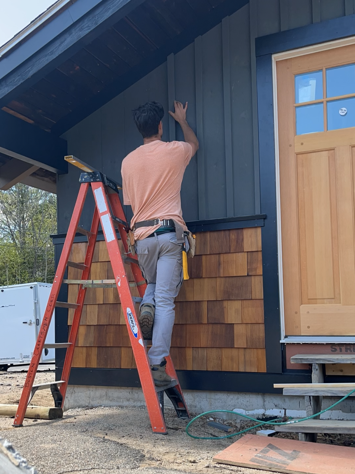 A man is standing on a red ladder, working on the exterior wall of a house, which has dark siding and a wooden door.