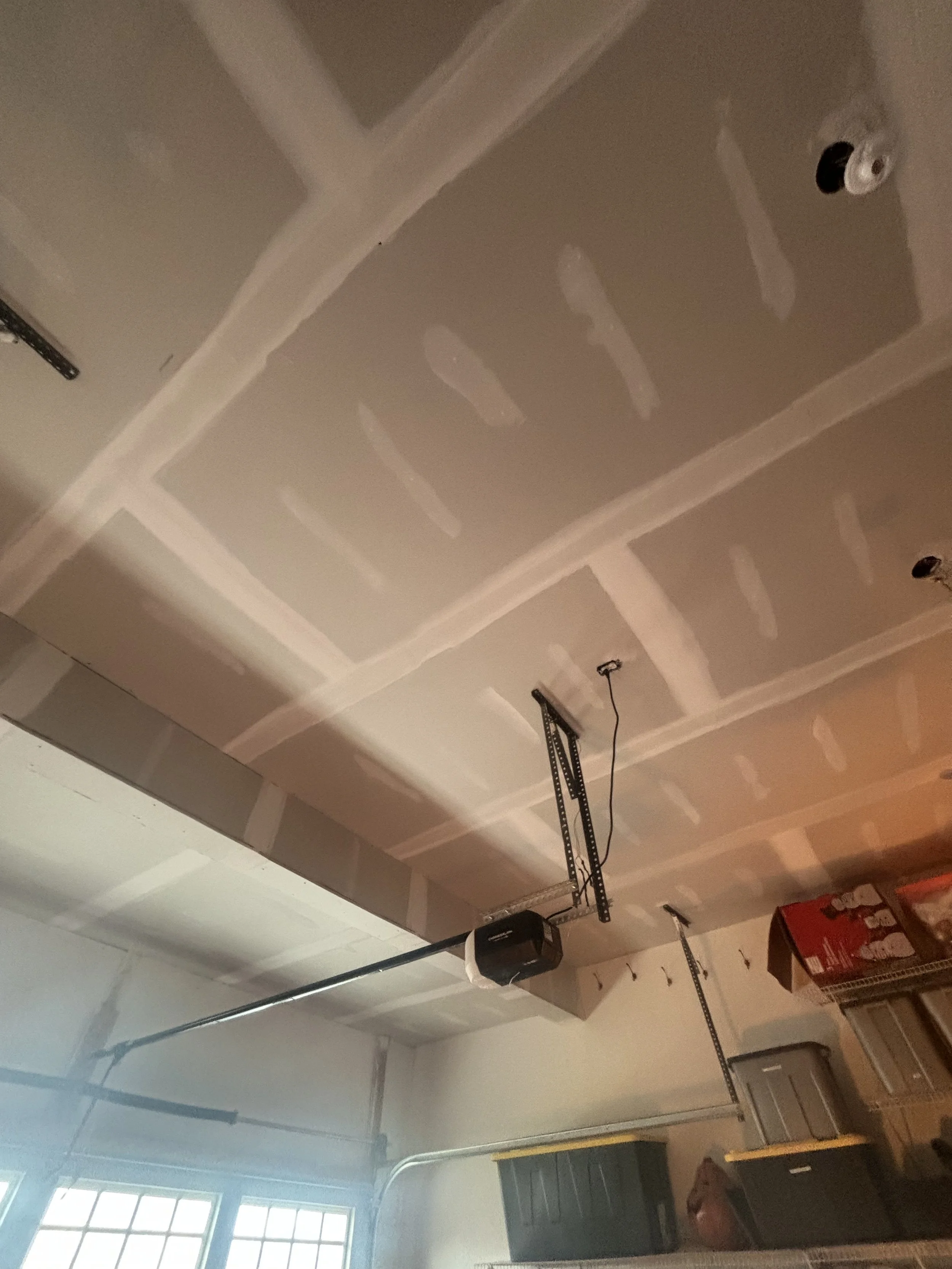 Interior view of a garage ceiling with drywall panels and electrical wiring, along with garage door openers and storage containers.
