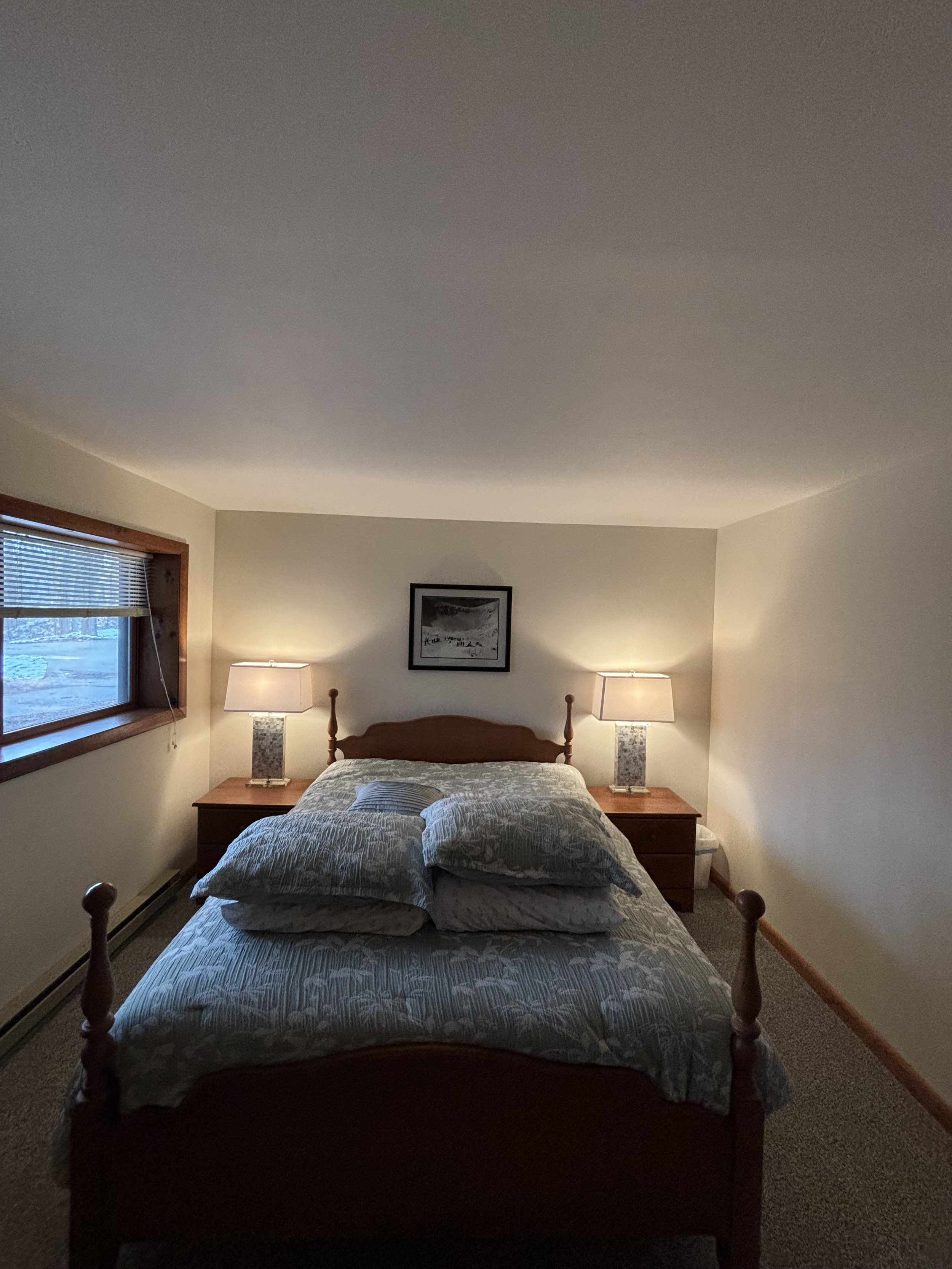 A cozy bedroom with a wooden bed frame and headboard, two bedside tables each with a lamp, a window with blinds on the left, a framed picture on the wall above the bed, and a pile of pillows and a comforter on the bed.