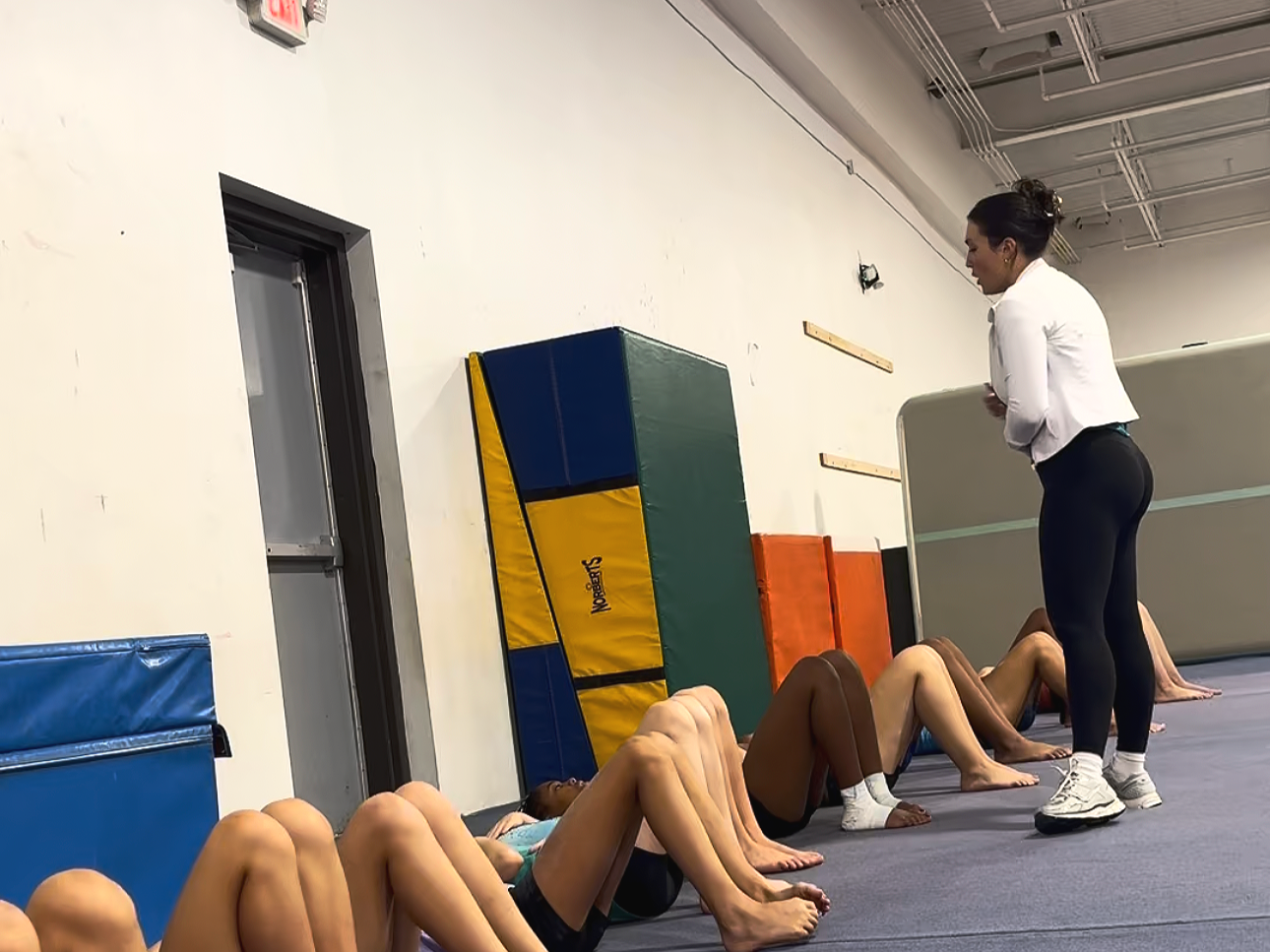 Instructor teaching a fitness class with participants doing sit-ups on the floor in a gym.