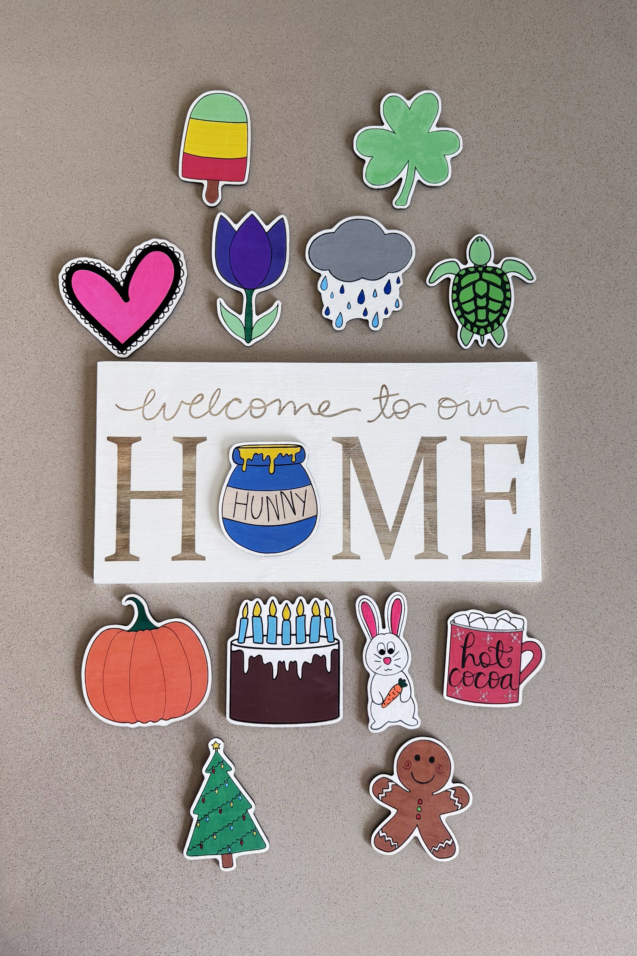 Decorative wood cutouts referred to as “Icons” arranged around a white sign that says 'welcome to our HOME,' with a jar labeled 'HUNNY' in place of the O in home. Additional "Icons" include a popsicle, tulip, pumpkin, hot cocoa cup, turtle, and more.