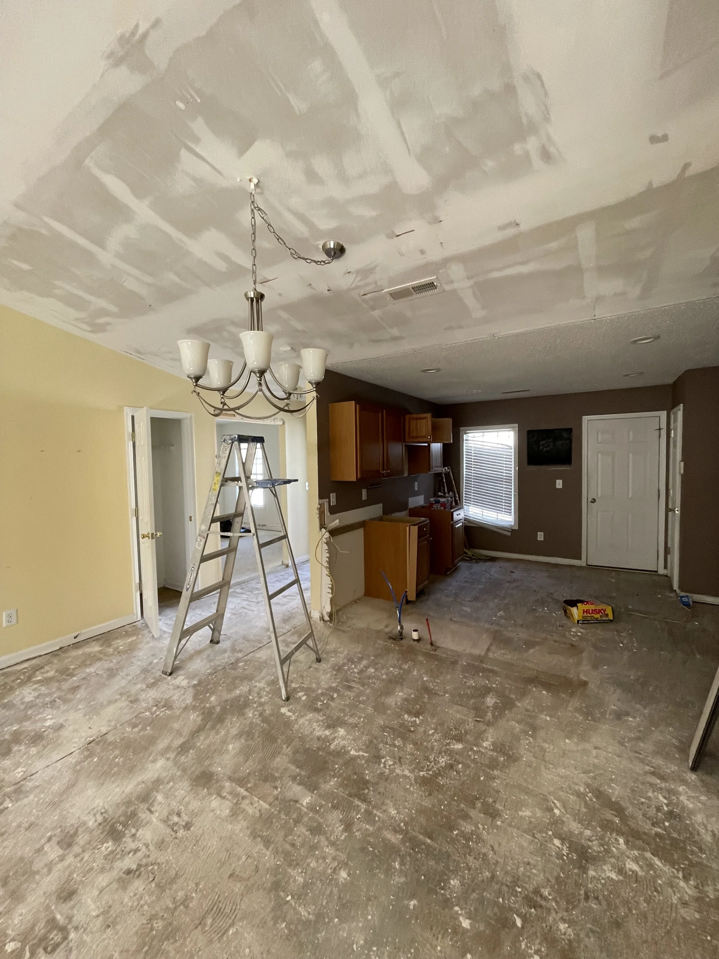Interior of a house under renovation with unfinished drywall ceiling and floor, ladder, and construction tools scattered around.