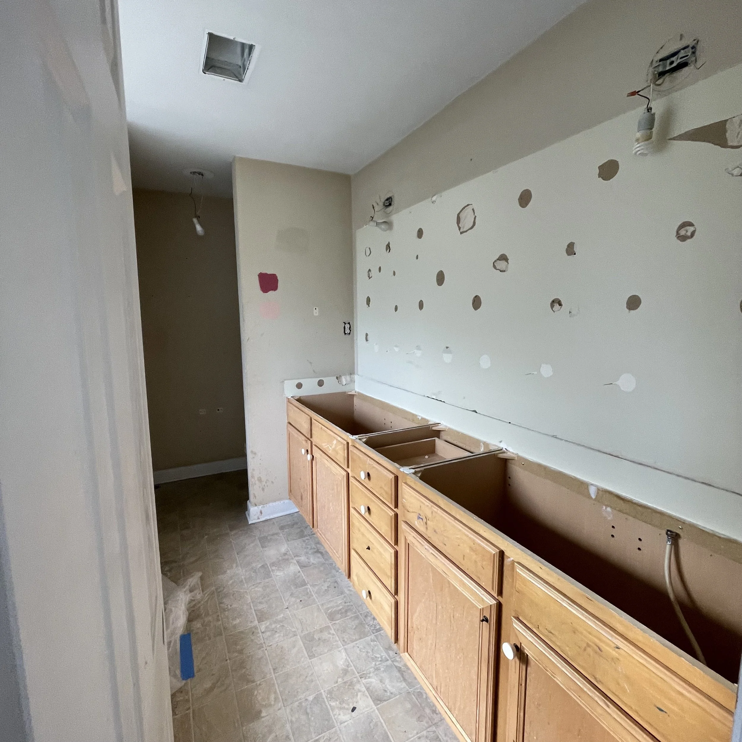 A room under renovation with wooden cabinets installed along one wall but no countertops. The walls are unfinished with holes and patches. There are two ceiling-mounted light fixtures without covers, and an air vent in the ceiling. Construction tools