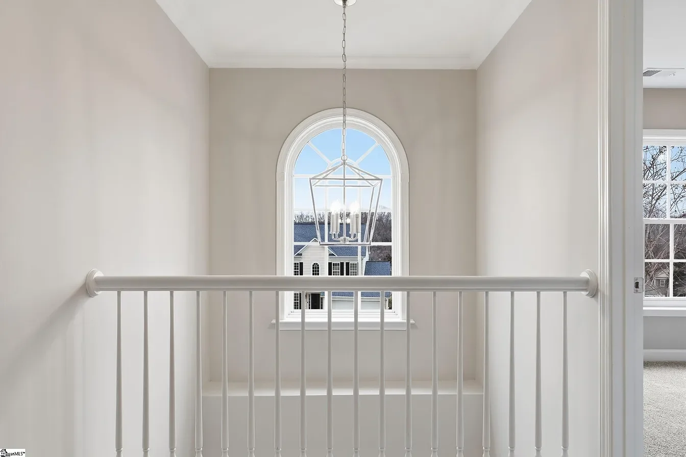 A bright, empty white-walled room with a large arched window in the center. A modern chandelier hangs from the ceiling. Part of a doorway with a white railing in the foreground and an adjacent room with another window are visible.
