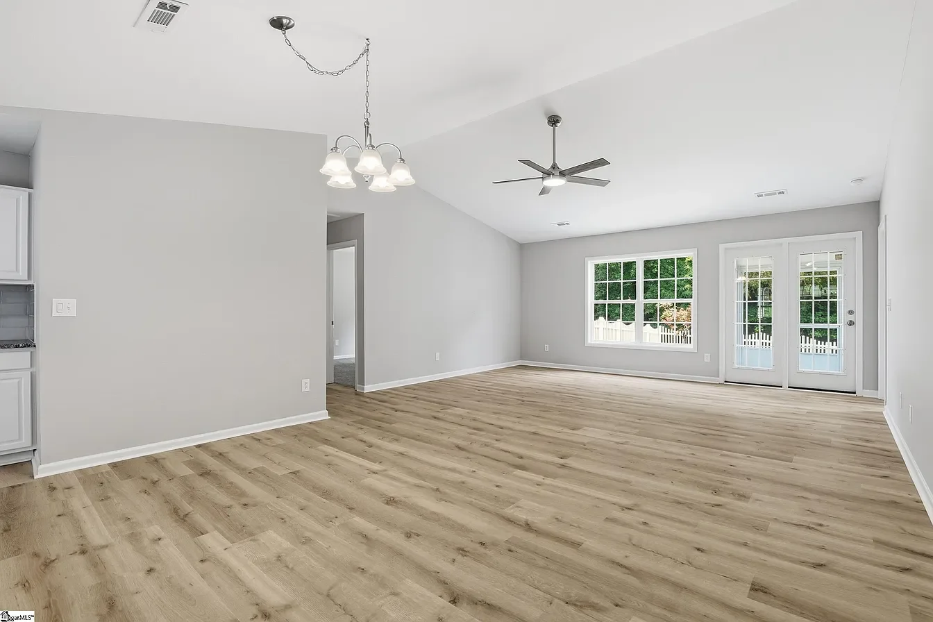Empty living room with large window and glass door, light wood flooring, white walls, ceiling fan, and chandelier, with a view of greenery outside.