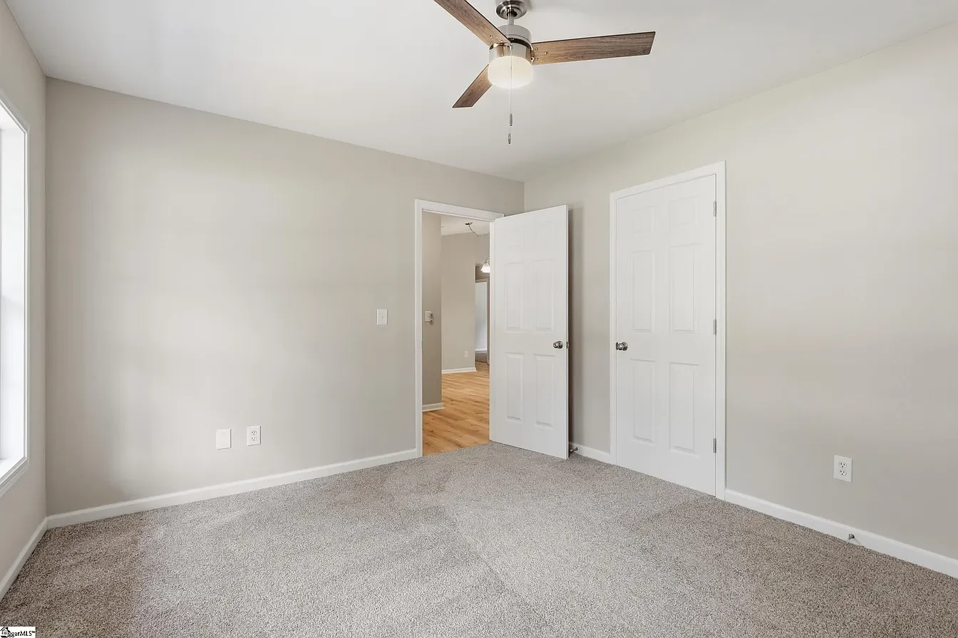 Empty bedroom with beige carpet, light gray walls, a ceiling fan with wooden blades, a large window on the left, and two white closet doors on the right.