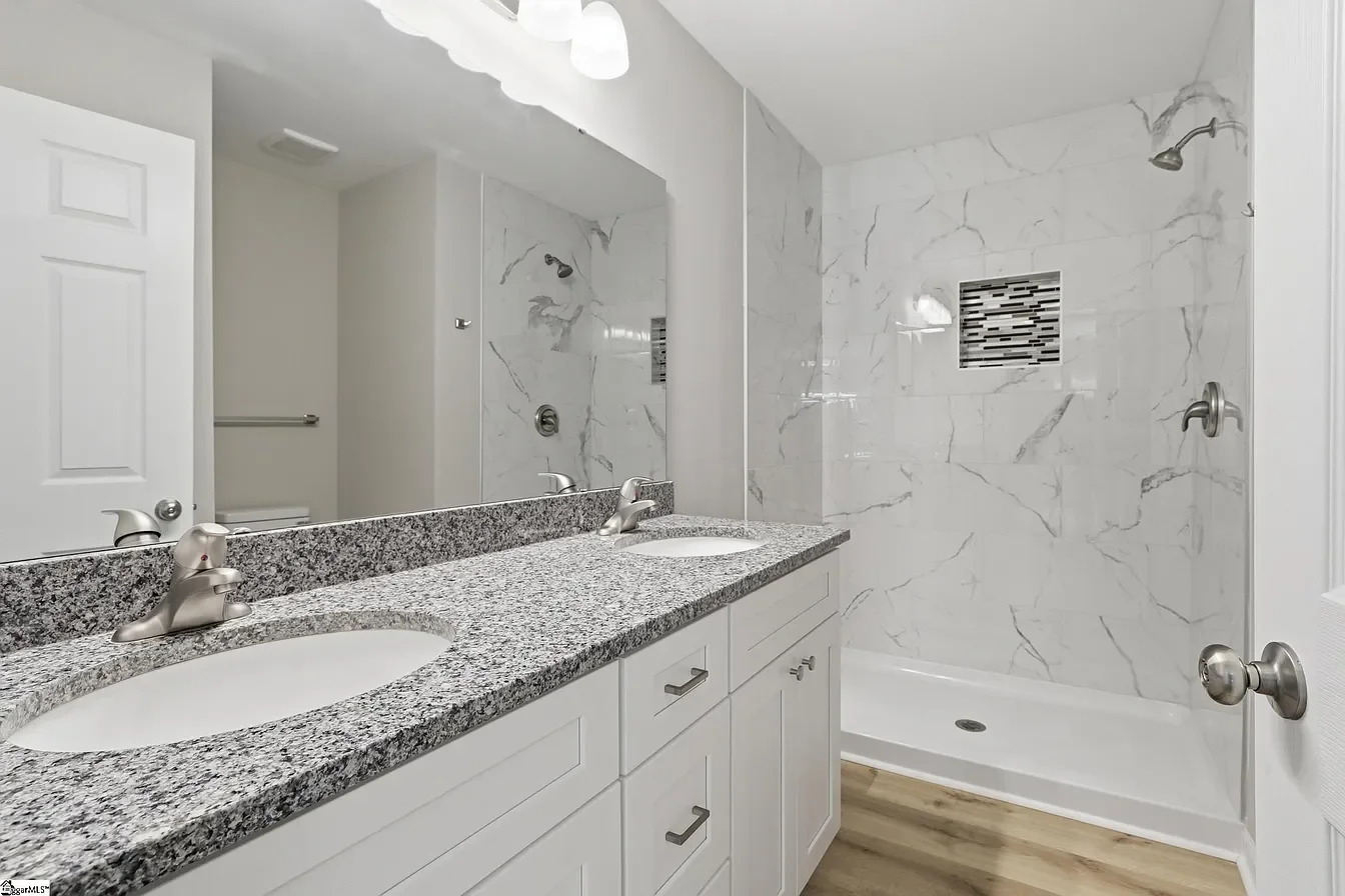 Modern bathroom with dual sink granite countertop, large mirror, marble shower with niche, and wood-look flooring.