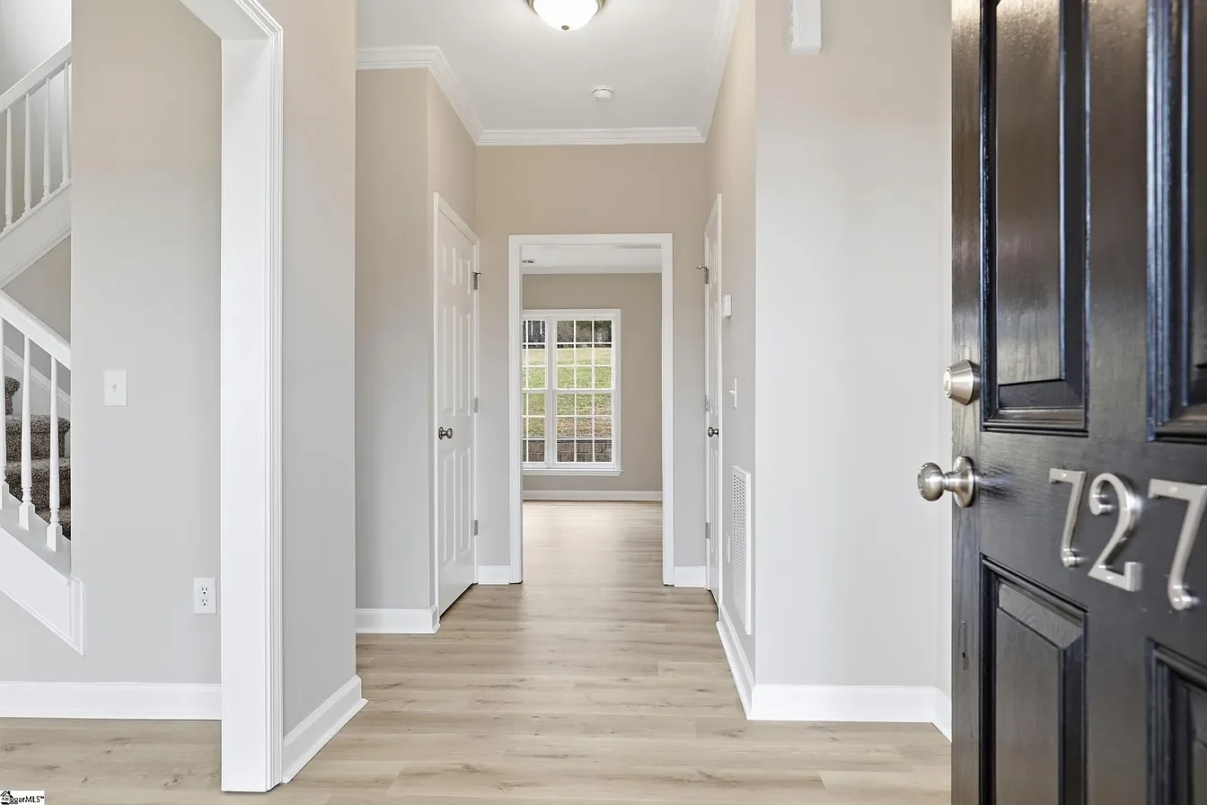 View of a modern house interior with a front door showing the house number 127. The entryway has hardwood floors, white trim, light gray walls, and doors leading to different rooms. There is a staircase on the left and a window at the back letting in