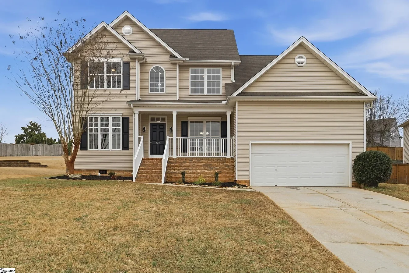 Front view of a two-story suburban house with beige siding, black shutters, a brick porch, and a two-car garage. The lawn is brownish, with a small tree on the left and a bush on the right.