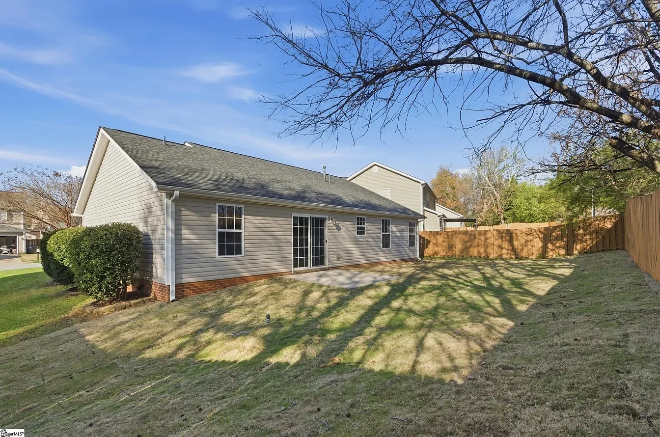 Backyard view of a house with beige siding, several windows, a sliding glass door, a small patio, a lawn with tree shadows, a wooden fence, and some bushes, under a blue sky.