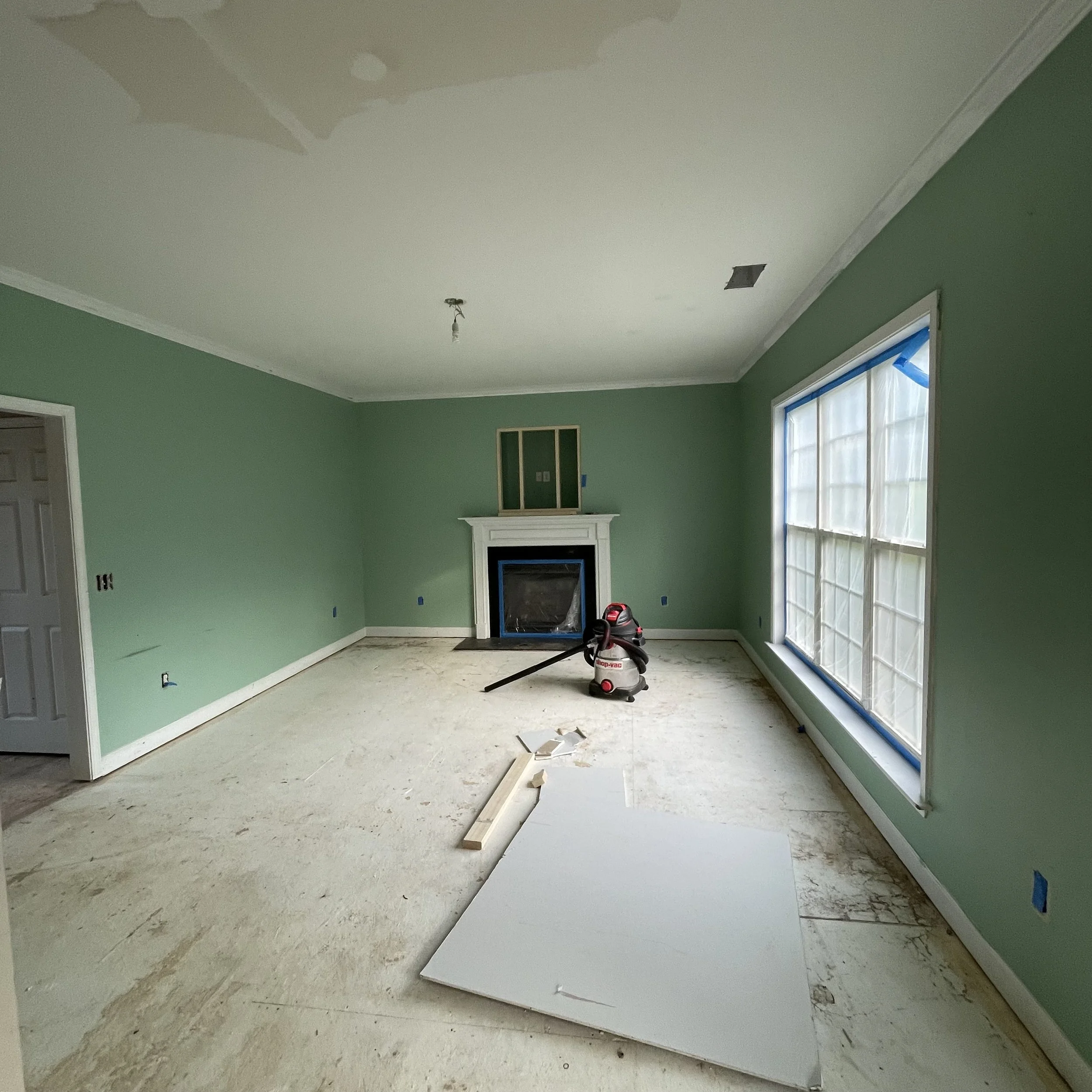 Interior of a room under renovation with green walls, a partially installed fireplace, a vacuum cleaner, and construction materials on the floor.
