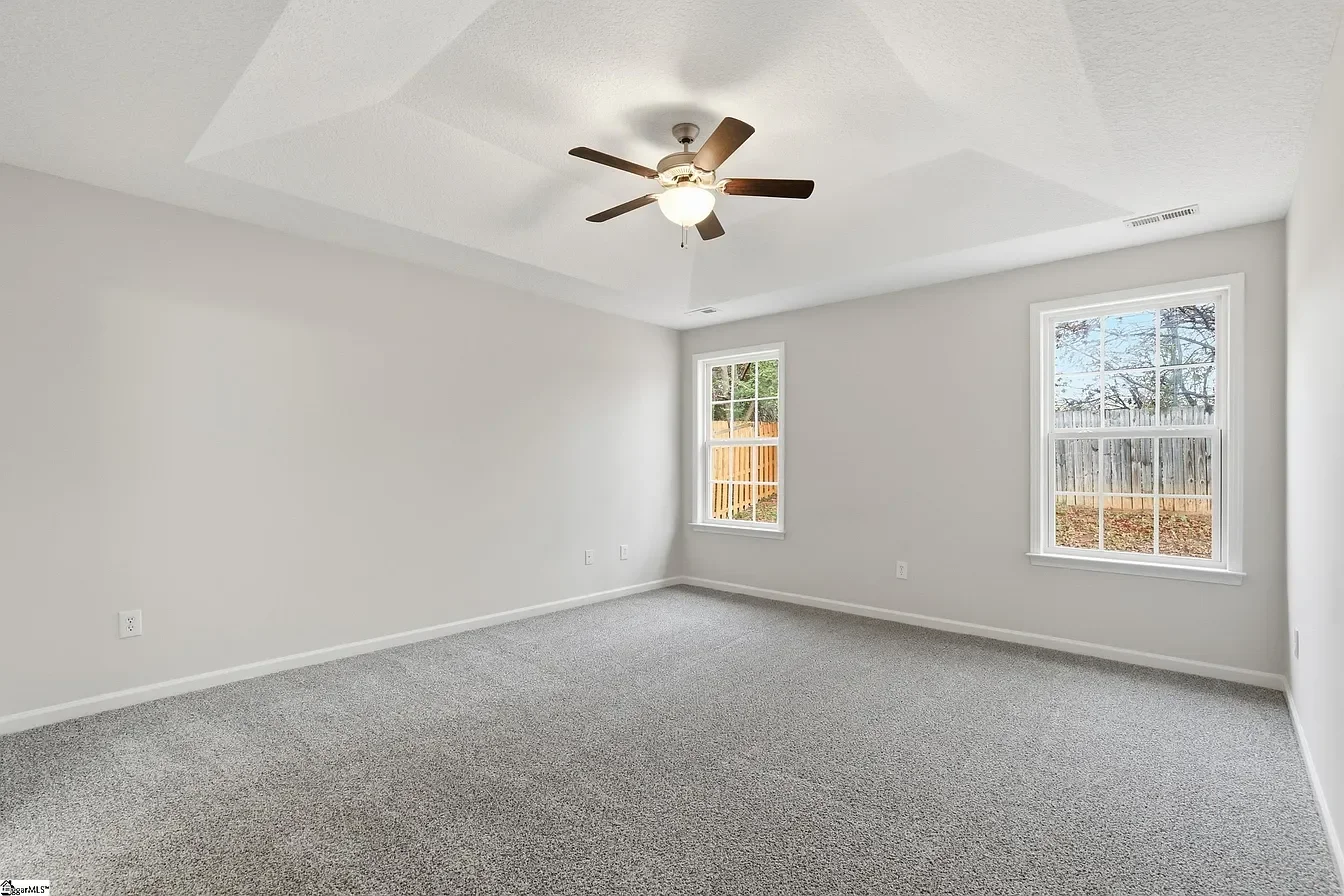 Empty room with gray carpet, white walls, two windows showing a fenced backyard, a ceiling fan with lights.