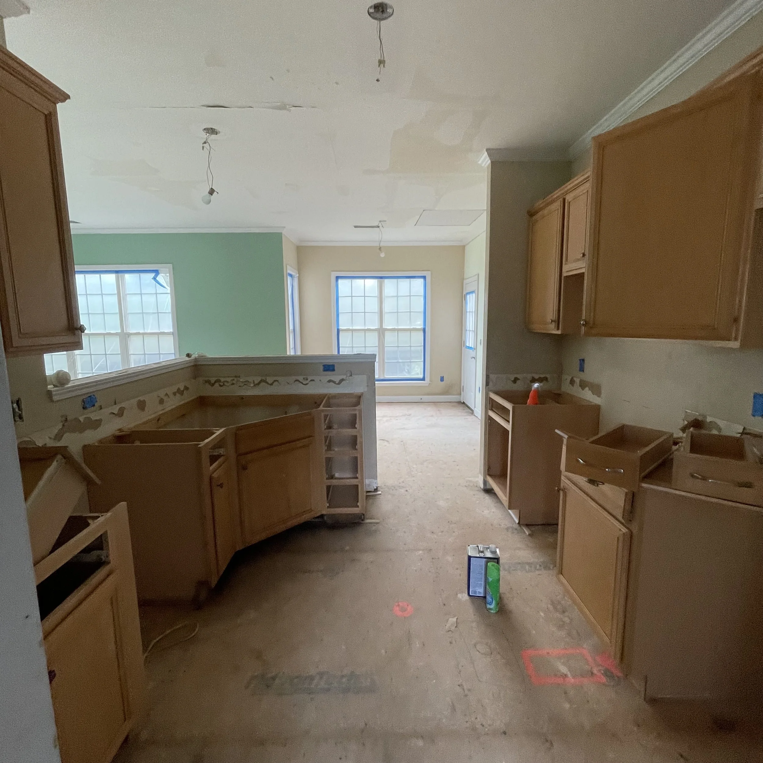 A room under renovation with cabinets installed, bare floors, and exposed wiring; windows are covered with blue painter's tape.