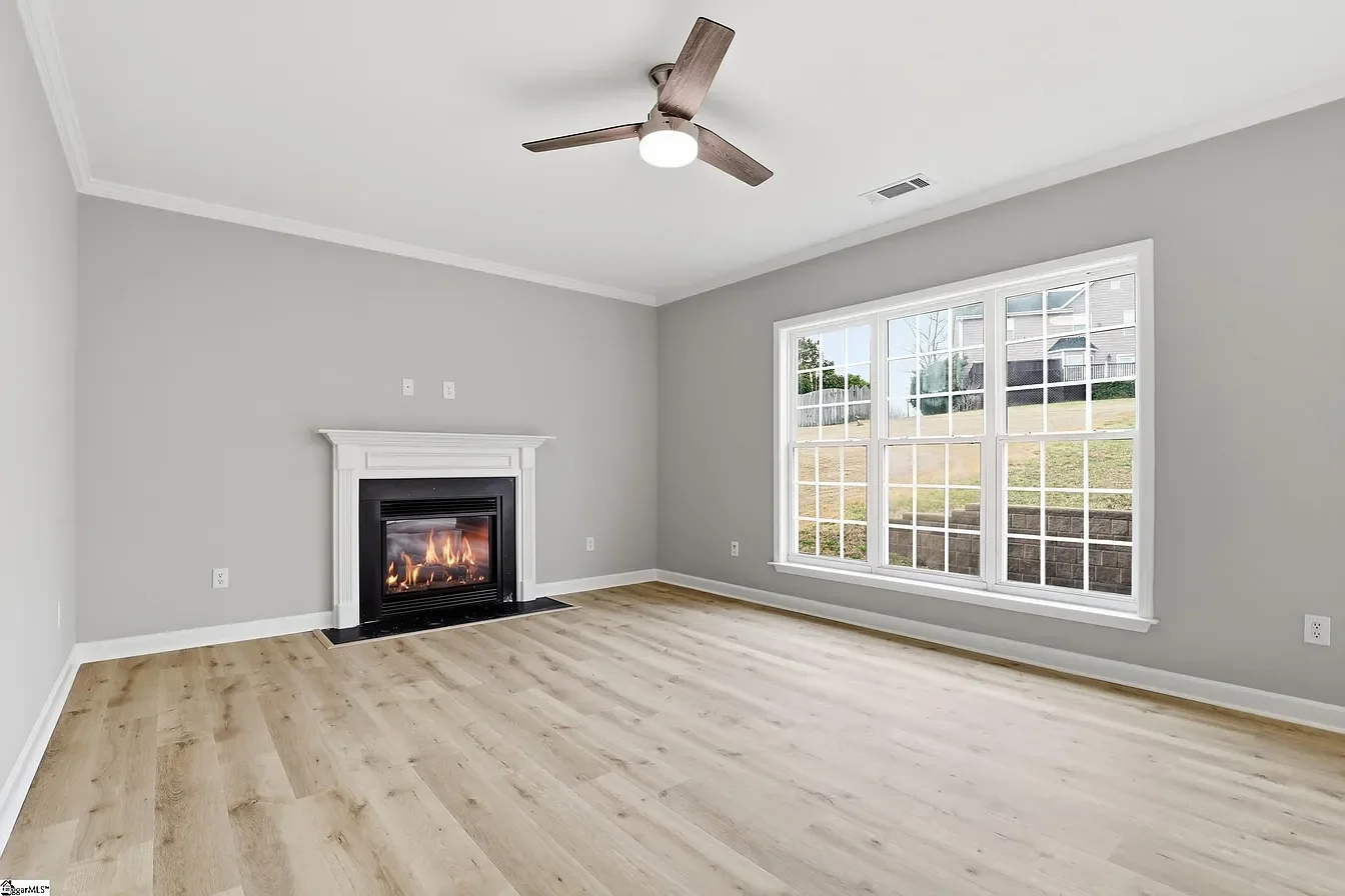 Empty living room with a fireplace, large window showing an outdoor grass yard, gray walls, wood laminate flooring, ceiling fan, and white crown molding.