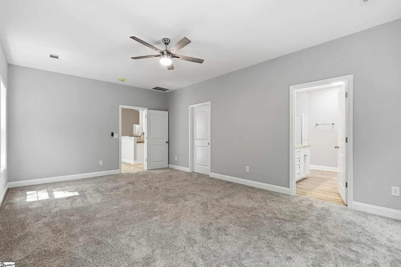 Empty room with gray walls, beige carpet, and three white doors, one leading to a bathroom, with ceiling fan and natural light.