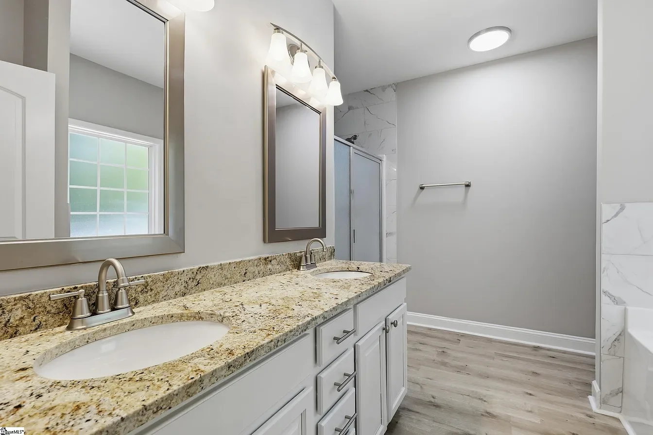 Modern bathroom with dual sinks, granite countertops, light gray cabinets, and large mirrors, with a window and a tiled shower in the background.