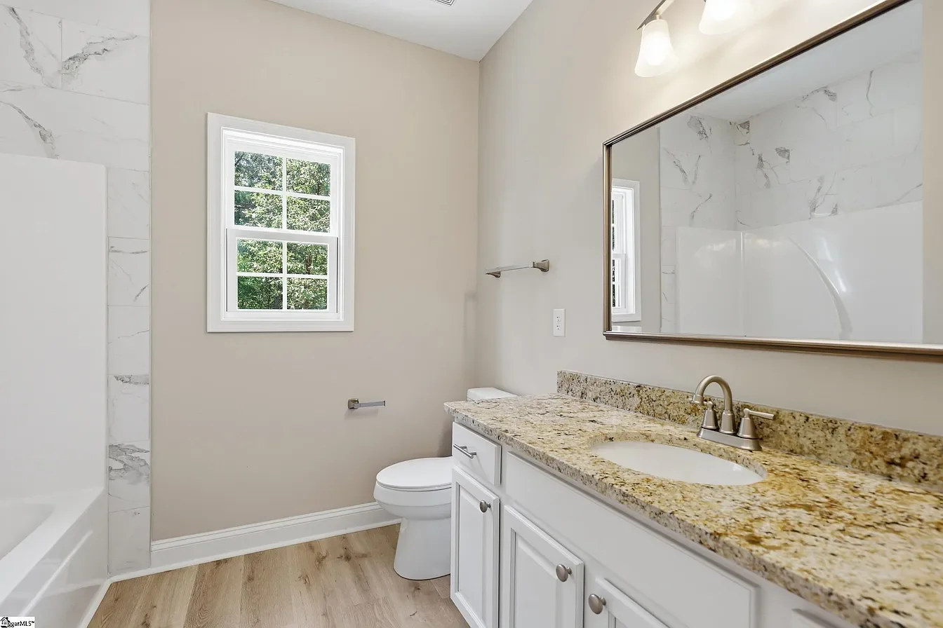 Bathroom with a granite countertop vanity, a large mirror, a window, a toilet, and a bathtub with marble tile surrounds.