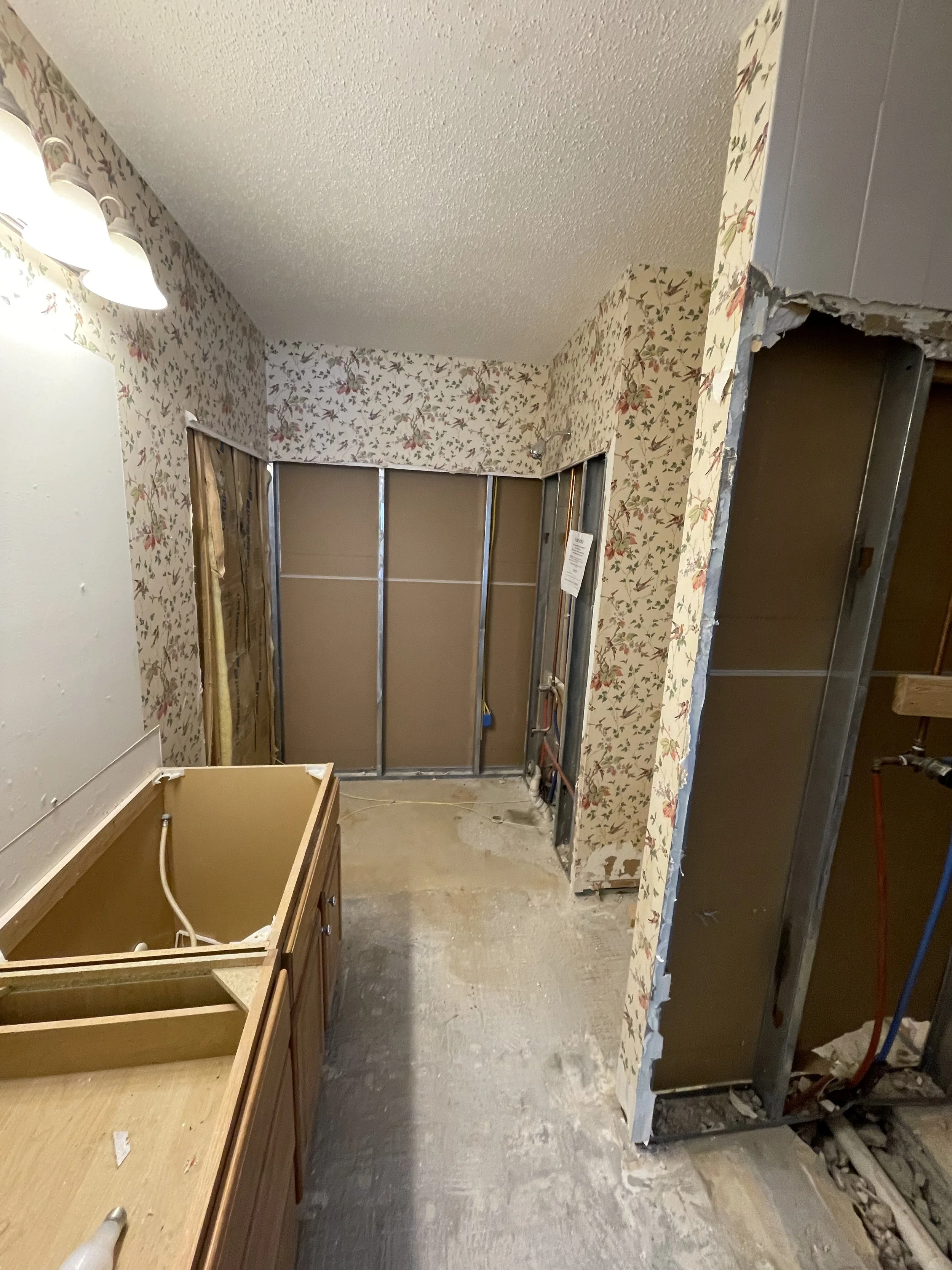 A room under renovation with exposed wall studs, a partially removed cabinet, and wallpaper with a floral pattern on the walls.